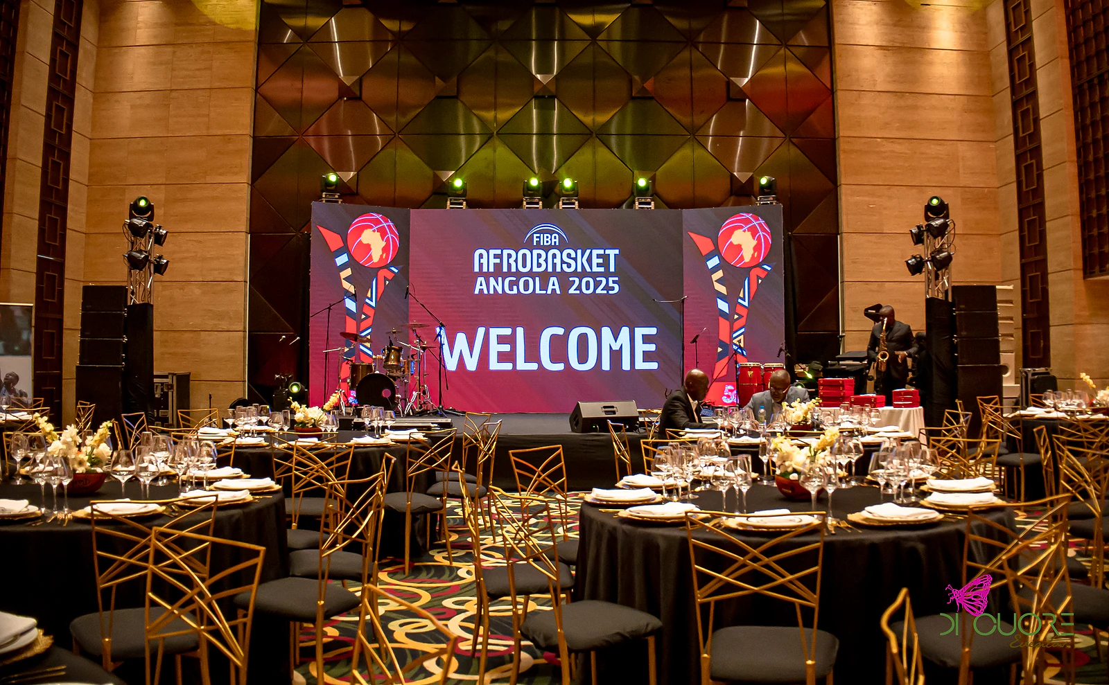 Sala de banquete com mesas redondas de toalha preta e cadeiras douradas, diante de um grande ecrã com a mensagem «Welcome» e o logótipo do FIBA AfroBasket Angola 2025