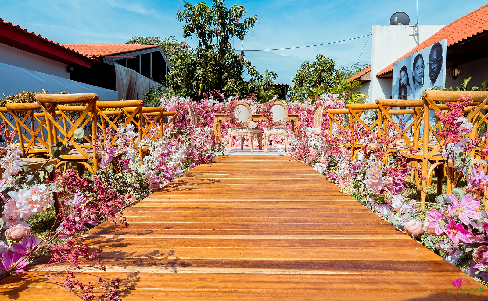 Passadiço de madeira entre flores cor-de-rosa e lilás e cadeiras de costas em X, até uma mesa de cerimónia ao fundo