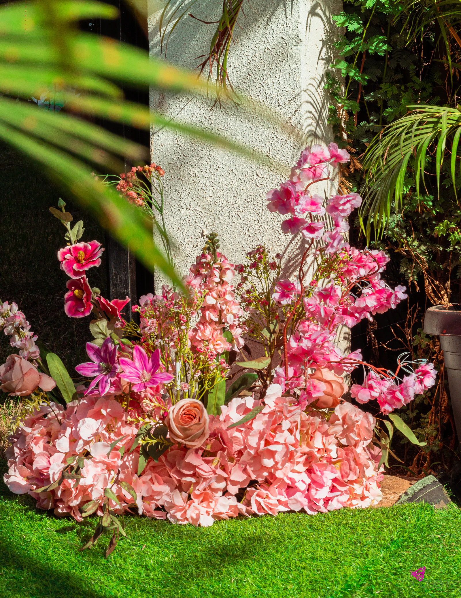 Maciço de hortênsias e flores cor-de-rosa ao pé de uma coluna branca, sobre relva, com uma folha de palmeira em primeiro plano