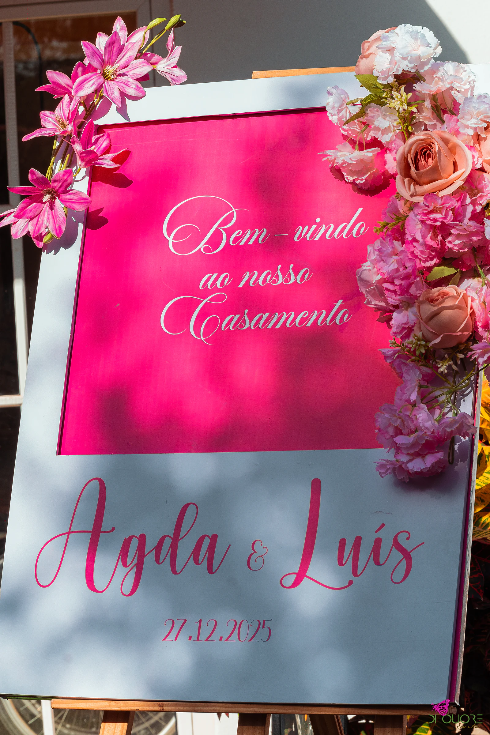 Cartaz de boas-vindas ao casamento, em fúcsia e cinzento, com os nomes Agda & Luís e a data, ladeado de flores cor-de-rosa