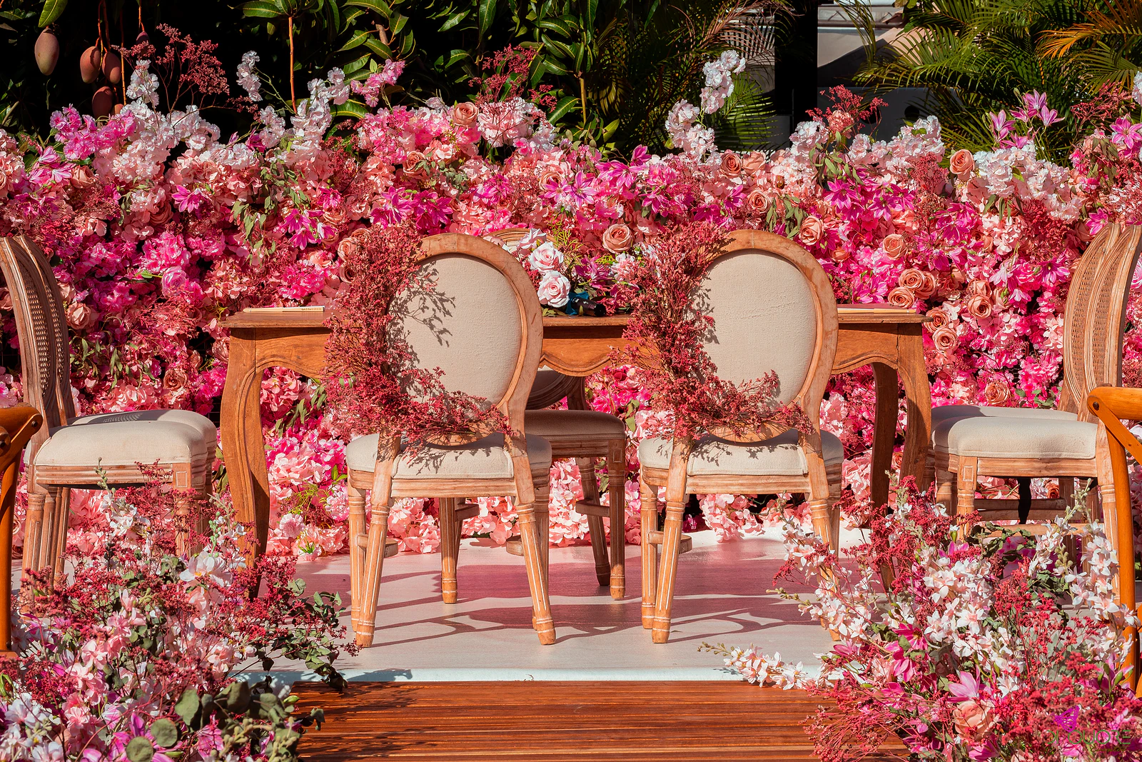 Mesa de madeira com cadeiras de costas ovais enfeitadas com grinaldas de flor miúda, diante de um painel de rosas e hortênsias cor-de-rosa, sobre um estrado de madeira