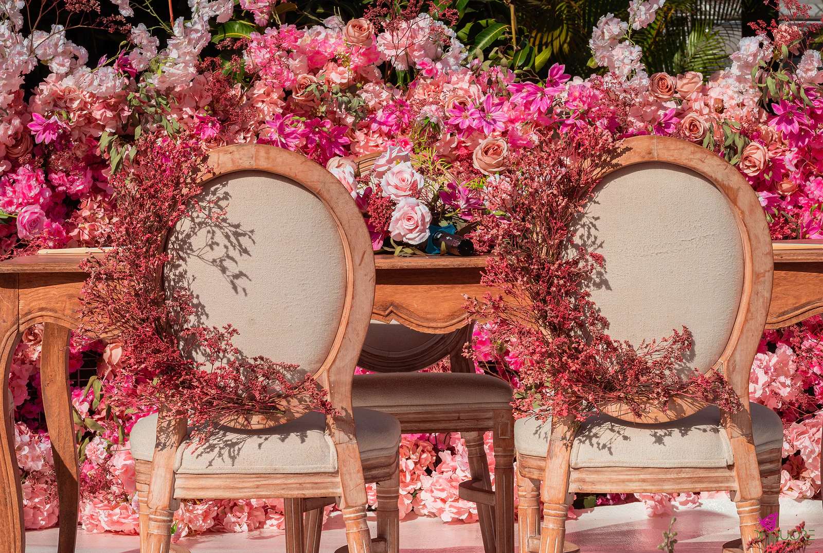 Duas cadeiras de costas ovais com grinaldas de flor miúda em tons de vinho, junto a uma mesa de madeira e a um painel de rosas cor-de-rosa