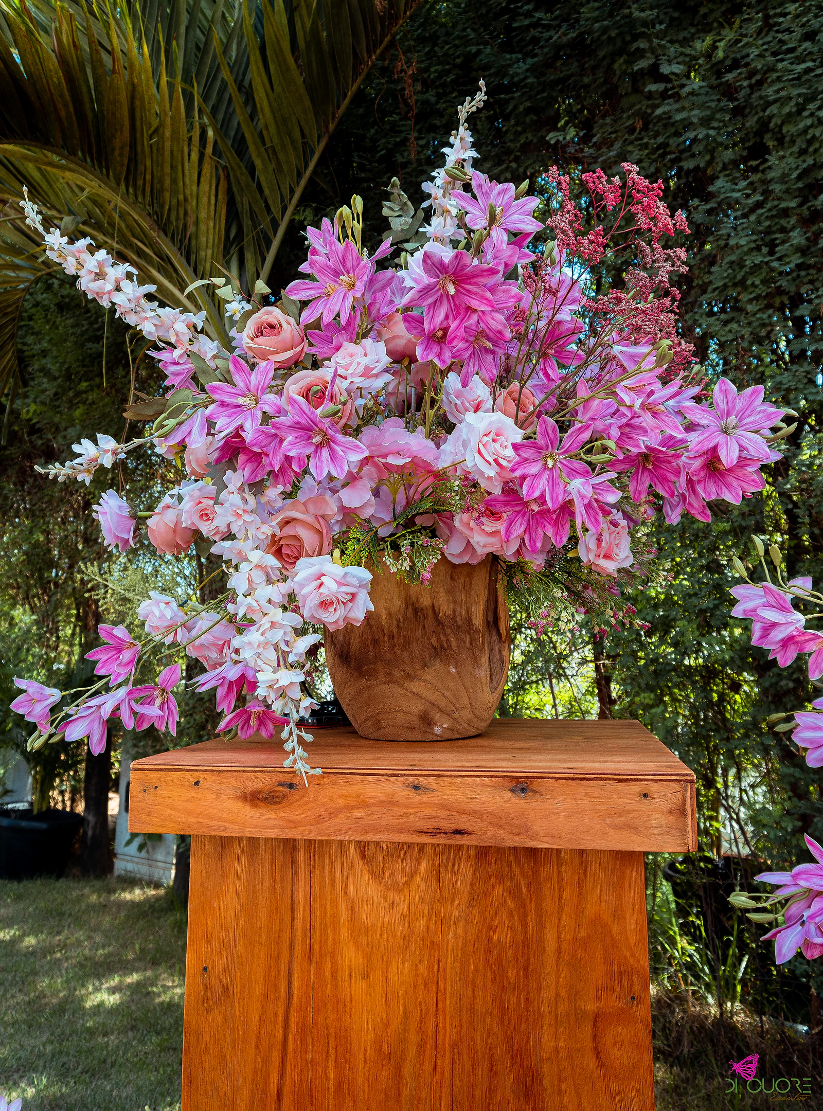 Arranjo alto de flores cor-de-rosa e brancas num vaso de madeira sobre um pedestal, com palmeira e folhagem ao fundo