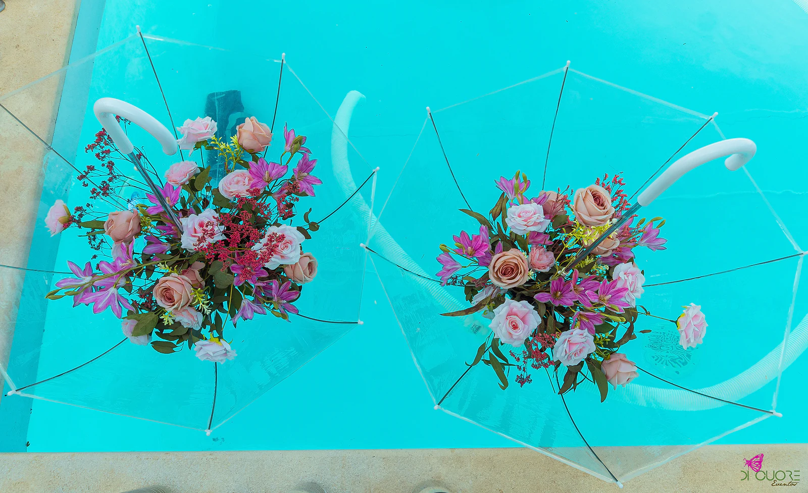 Dois guarda-chuvas transparentes com ramos de rosas e flores lilás, vistos de cima sobre a água turquesa da piscina