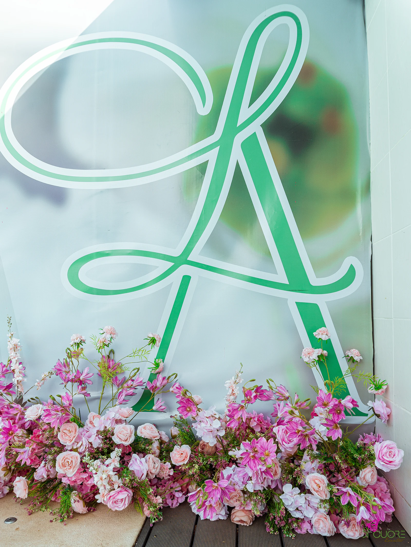 Monograma A em verde sobre um painel branco, com flores cor-de-rosa e brancas ao fundo do painel