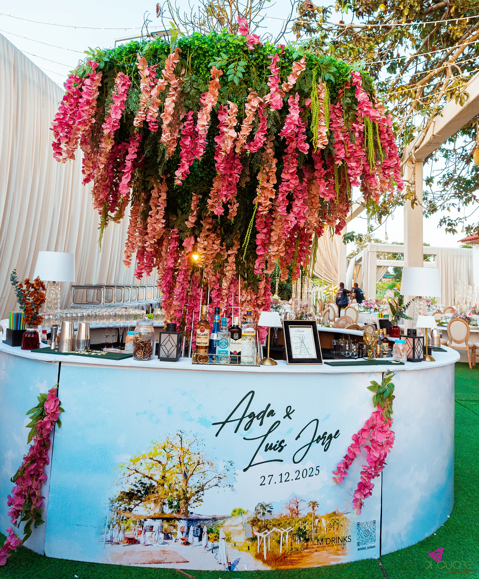 Bar redondo pintado com os nomes dos noivos, sob uma copa de flores suspensas em rosa e coral e folhagem