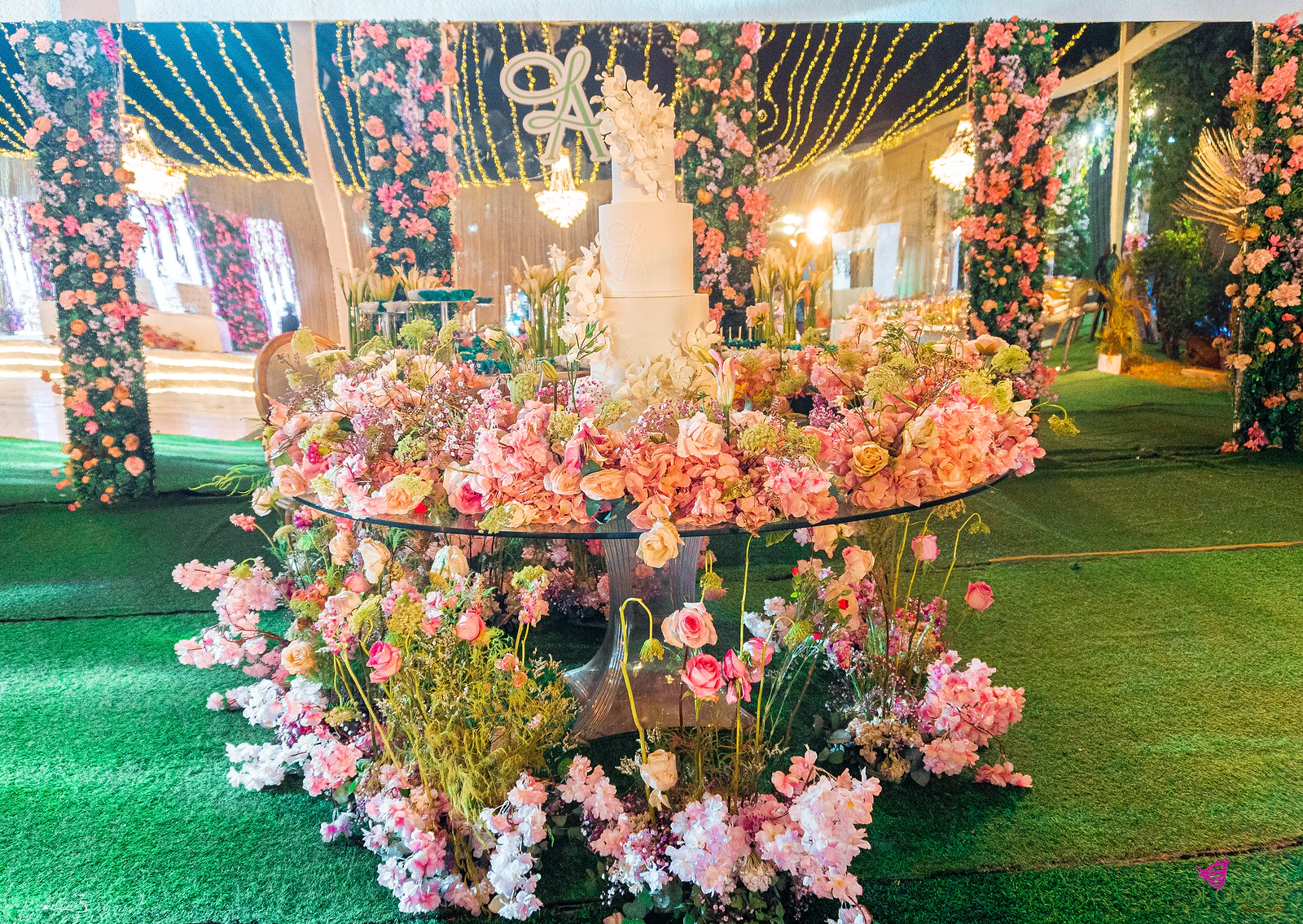 Mesa de vidro coberta de flores cor-de-rosa e pêssego com o bolo de vários andares, entre colunas floridas e cortinas de luzes