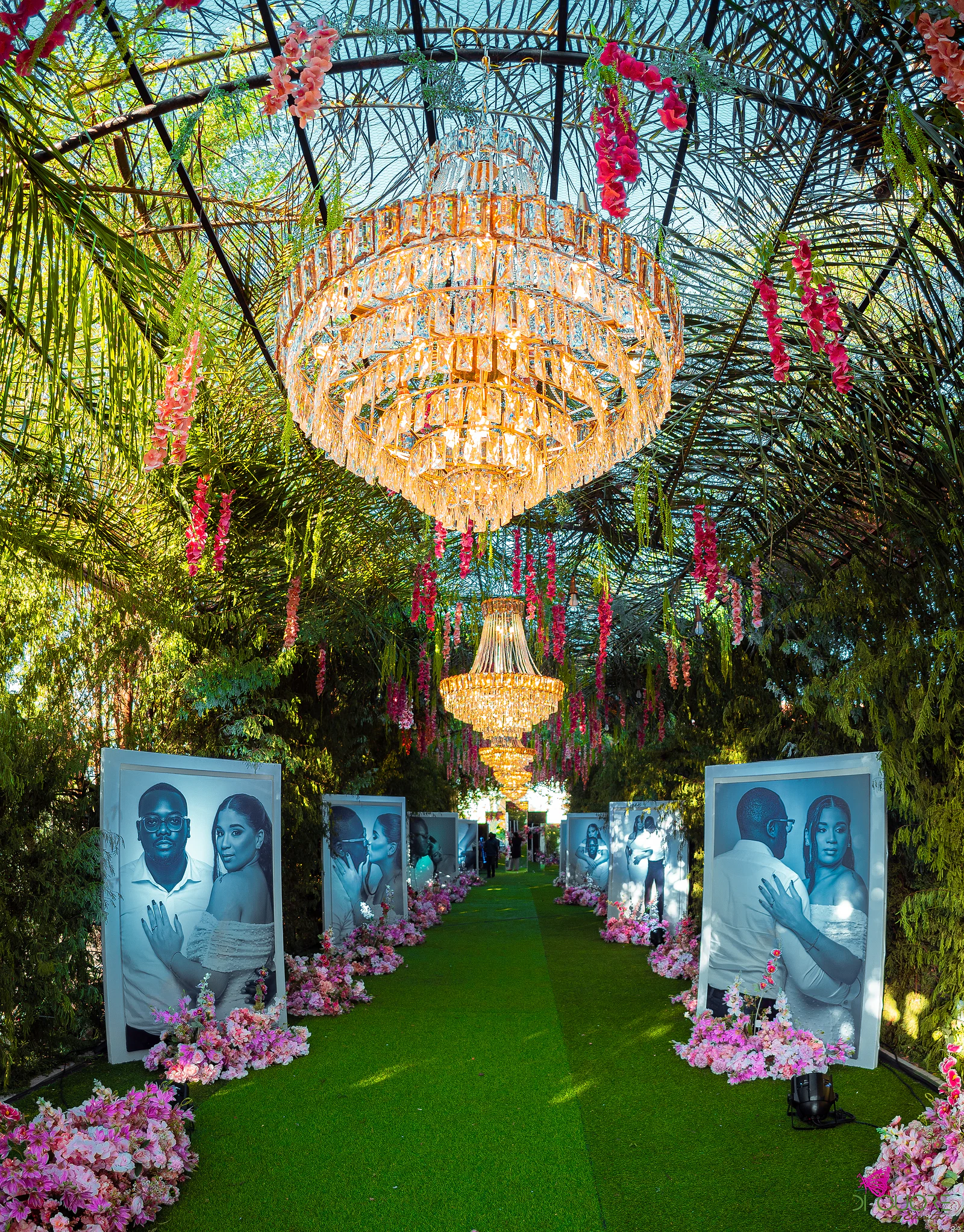 Corredor de relva sob um grande lustre de cristal e flores suspensas, ladeado de retratos dos noivos e flores cor-de-rosa