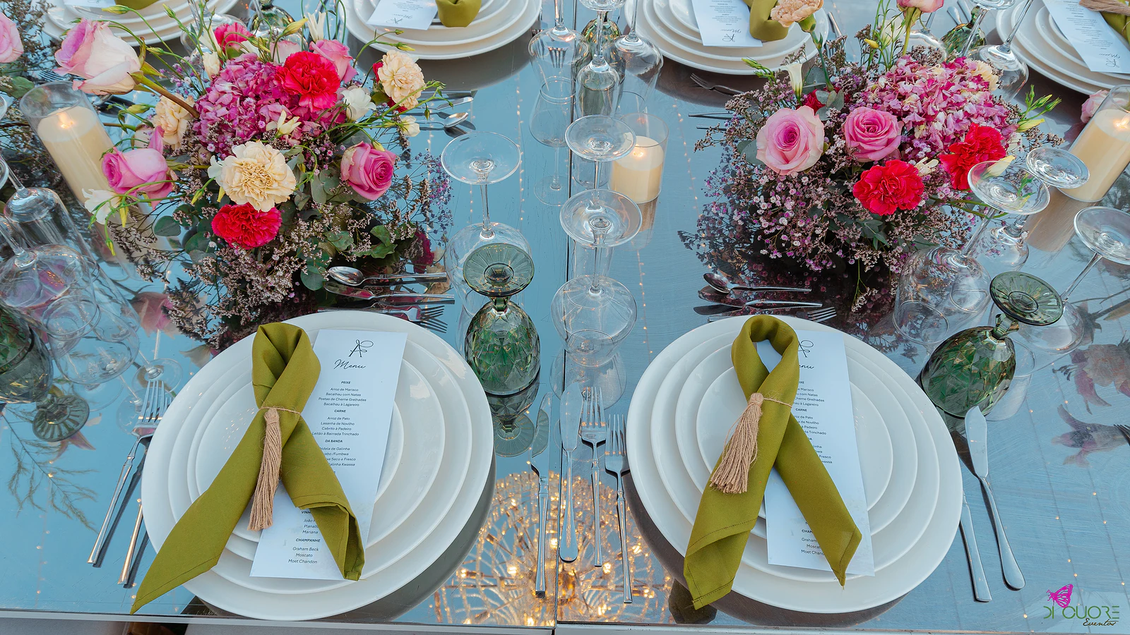 Mesa espelhada com pratos brancos, guardanapos verde-azeitona com borla e menu, e arranjos de rosas e cravos cor-de-rosa e vermelhos