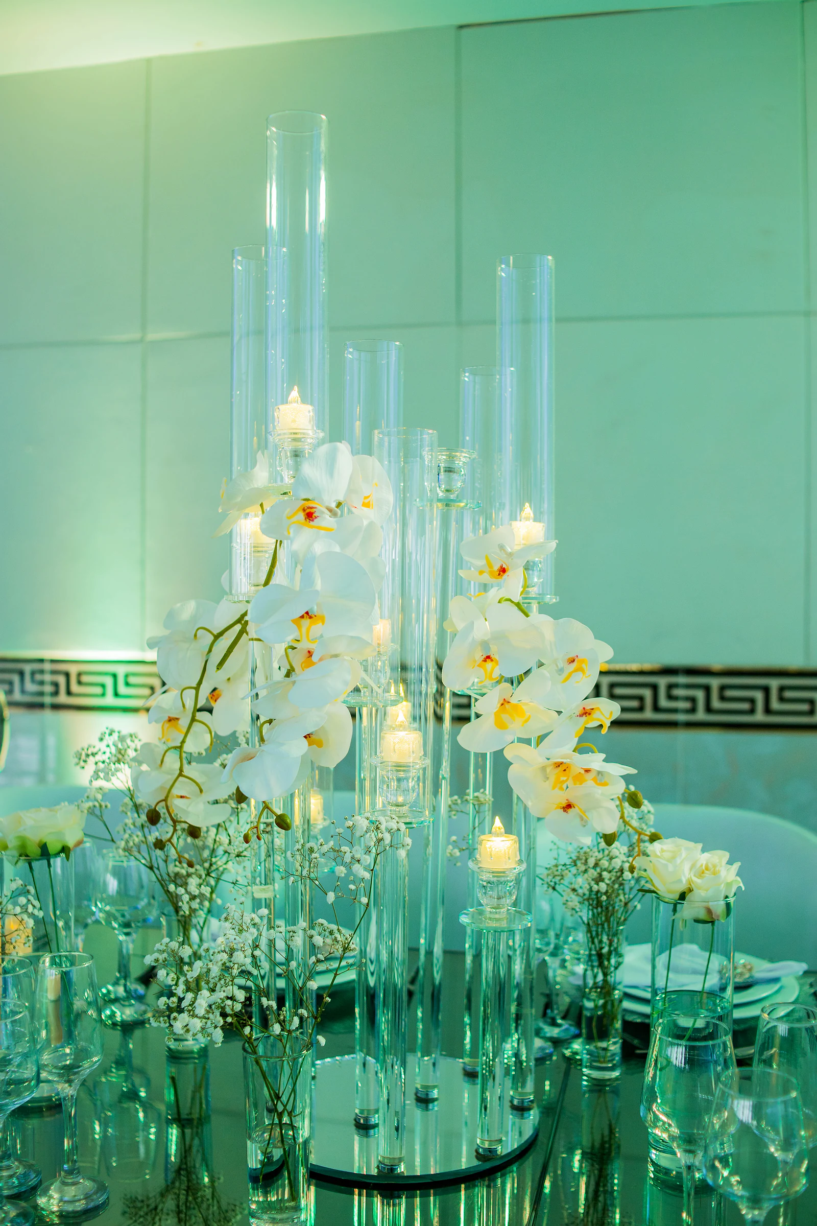 Centro de mesa com orquídeas brancas e velas em cilindros de vidro de alturas diferentes sobre um espelho