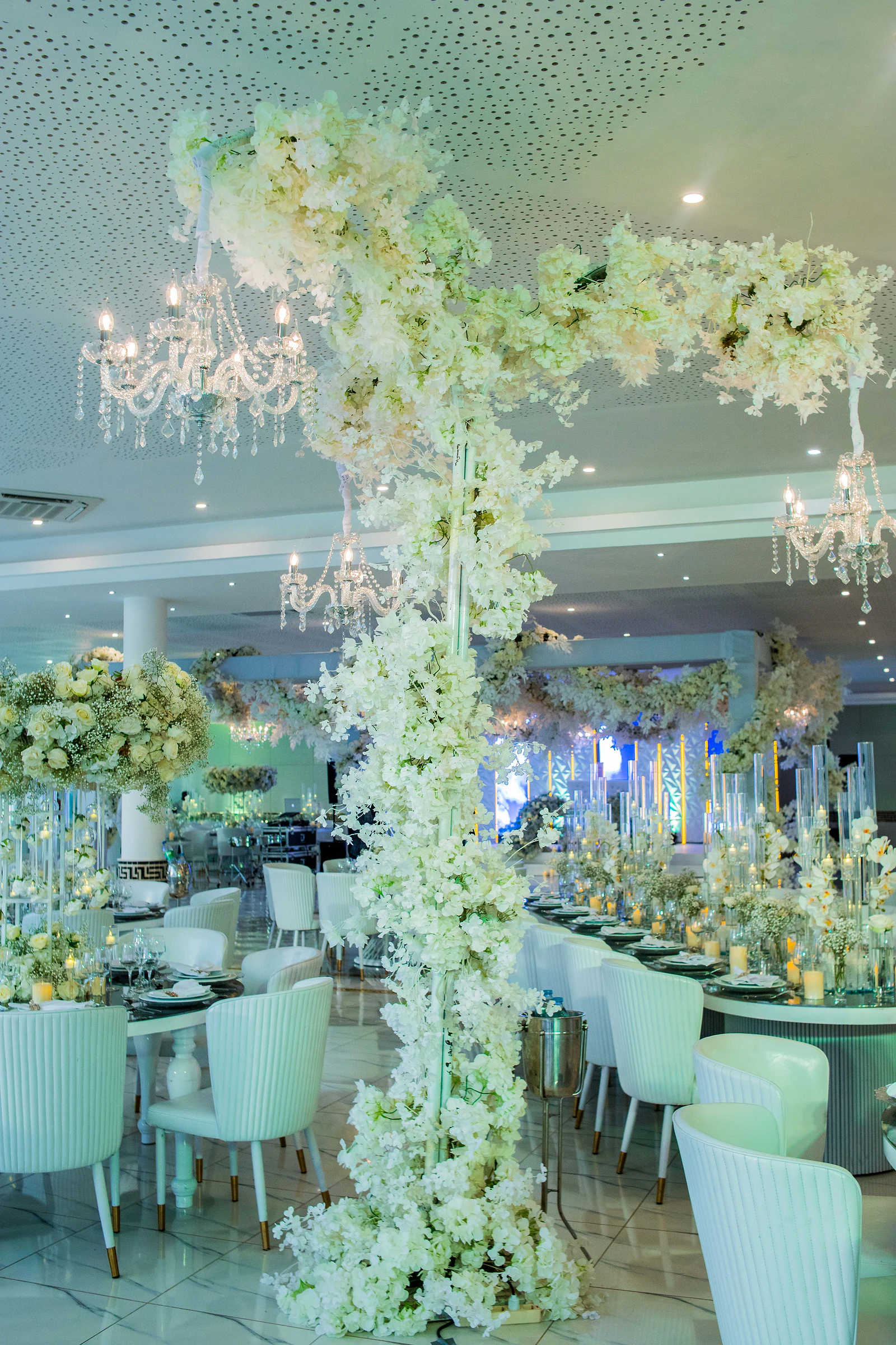 Coluna de flores brancas com braço em arco e candelabros de cristal suspensos, sobre mesas postas num salão de cadeiras brancas