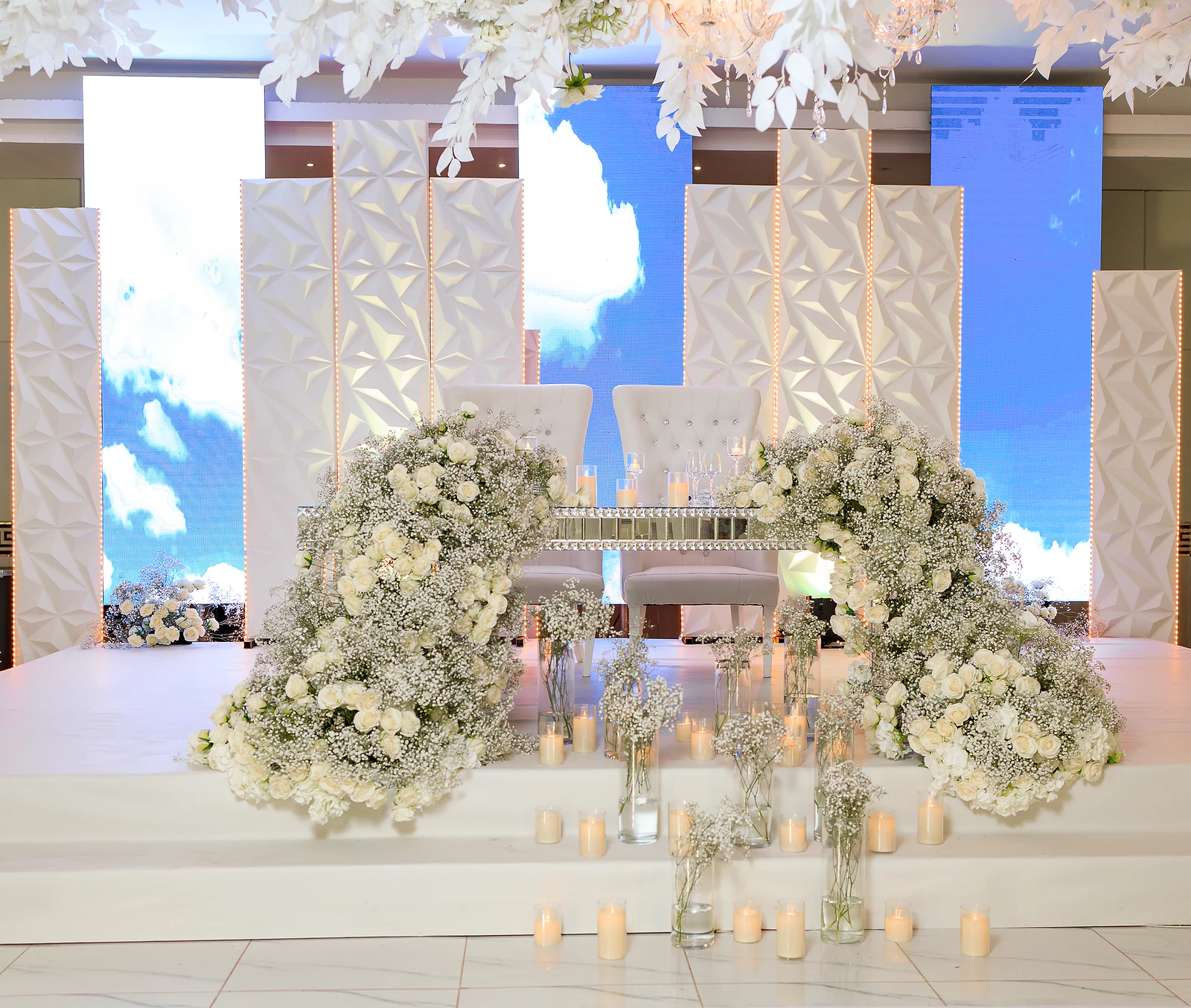 Mesa dos noivos com duas cadeiras brancas entre cascatas de rosas e gipsófila, velas no chão e painéis brancos com ecrãs de céu azul