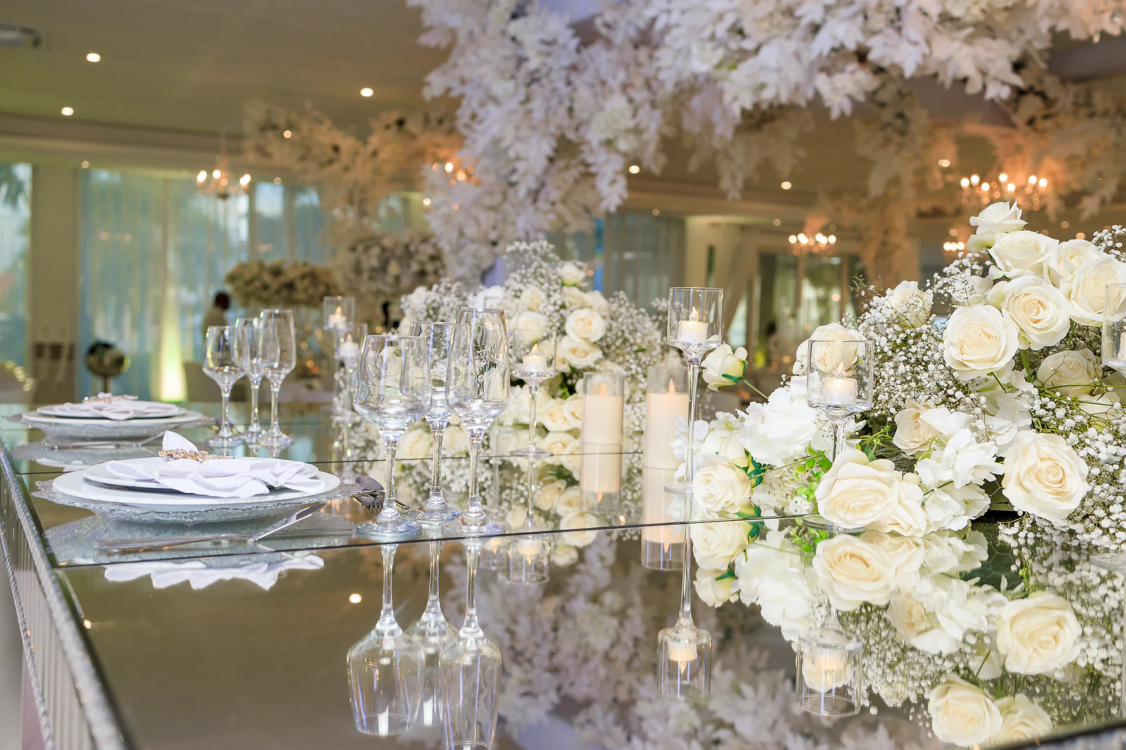 Mesa de espelho com ramos de rosas brancas e gipsófila, velas e copos de cristal, com flores brancas suspensas ao fundo