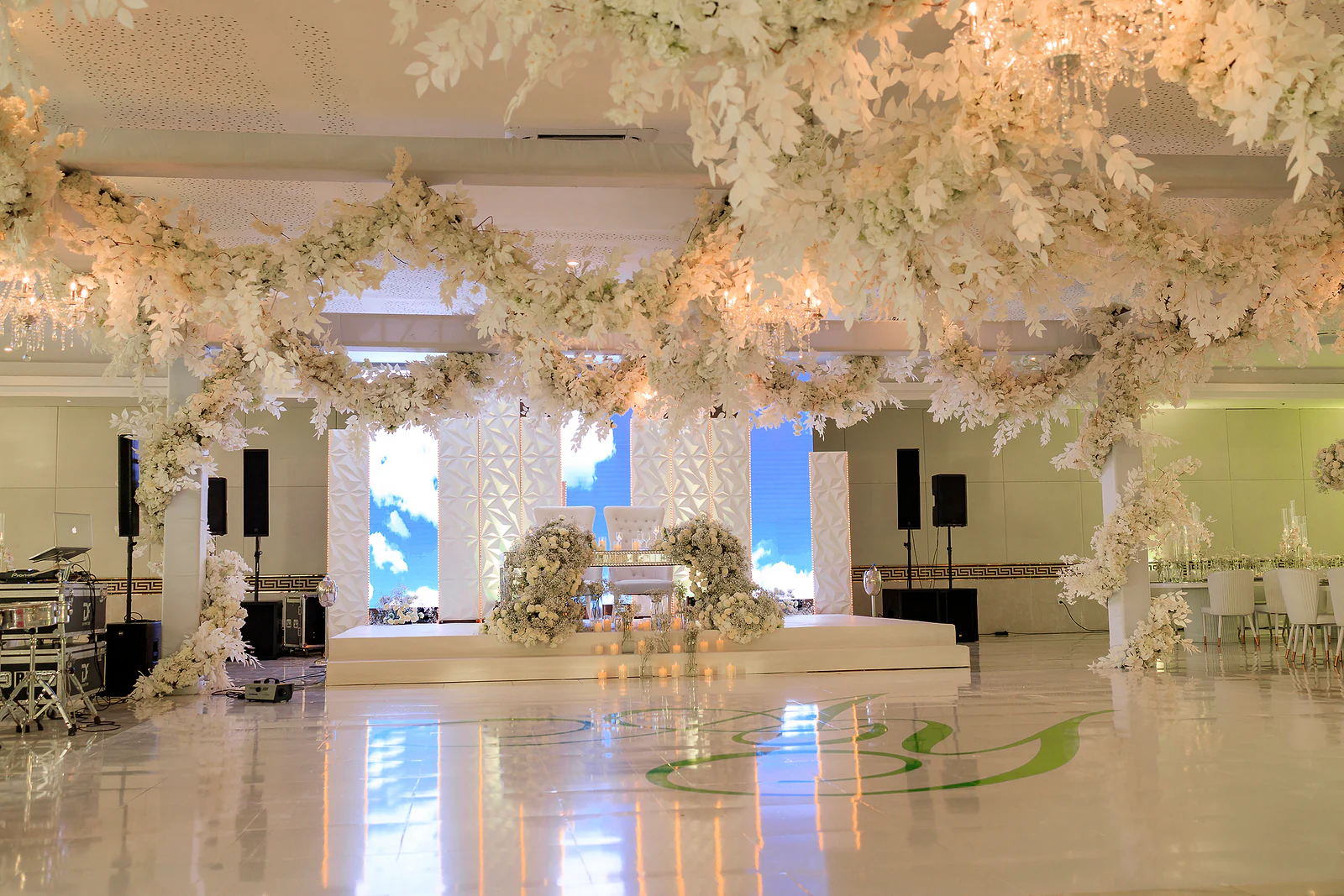 Salão com o tecto coberto de ramos de flores brancas, palco ao fundo com ecrã de céu azul e pista de dança brilhante