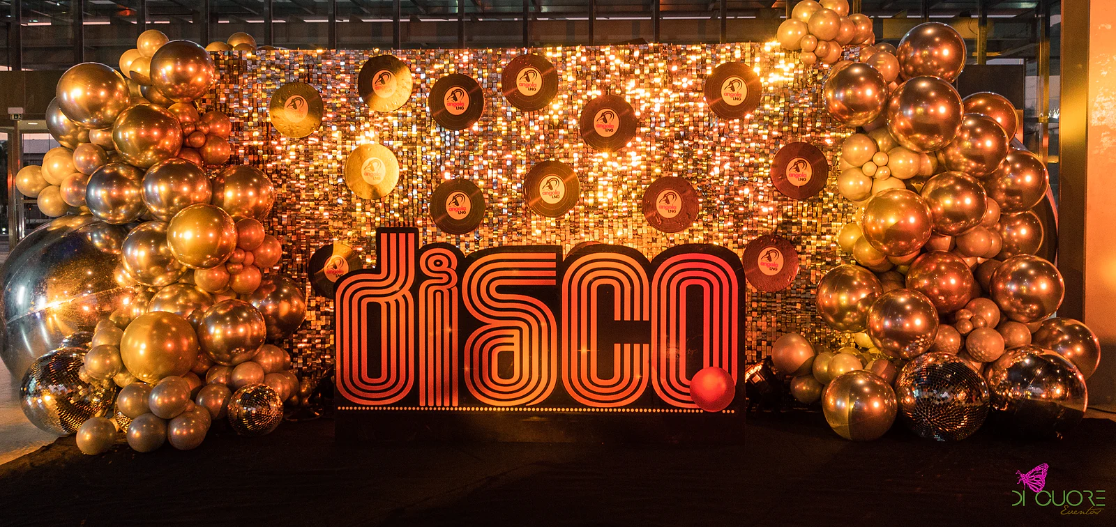 Gold sequin wall with vinyl records and the word disco in glowing red letters, flanked by clusters of gold and silver metallic balloons