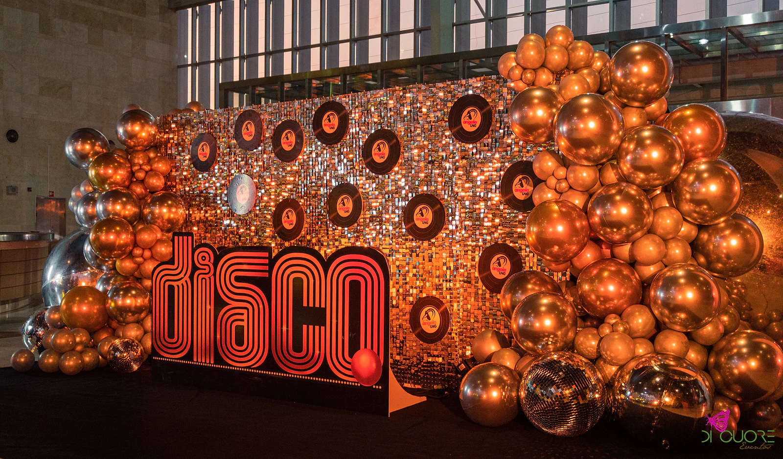 Painel de lantejoulas cor de cobre com discos de vinil, letreiro luminoso “disco” e balões cromados em cobre e prata