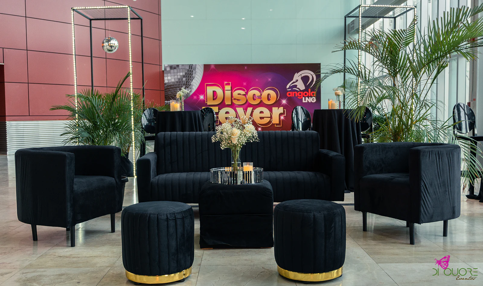 Lounge com sofá e cadeirões de veludo preto, pufes de base dourada e mesa de centro com rosas brancas, diante do painel “Disco Fever”