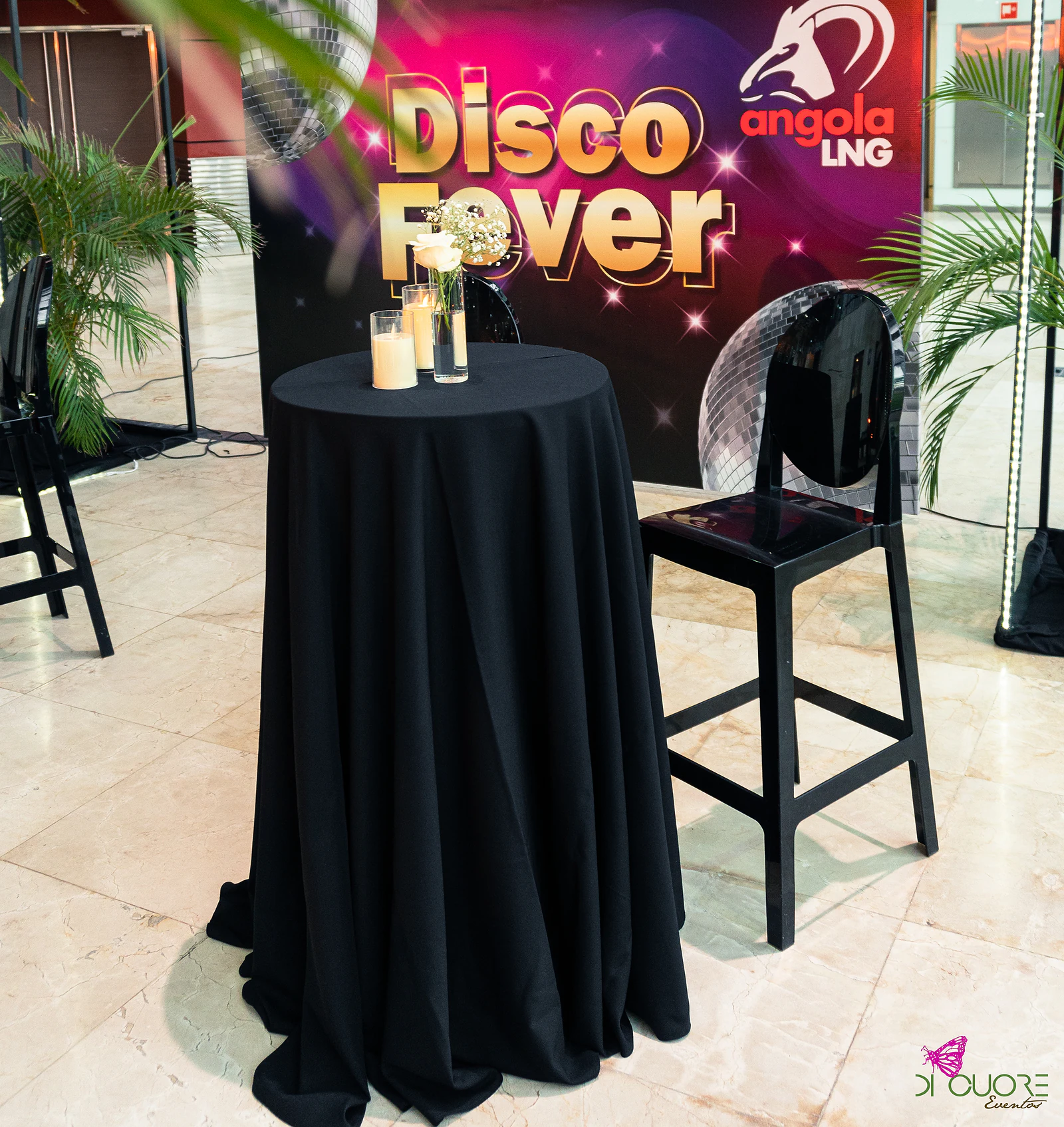 Mesa alta de toalha preta com velas e rosas brancas, diante do painel “Disco Fever” da Angola LNG, junto a um banco preto e palmeiras