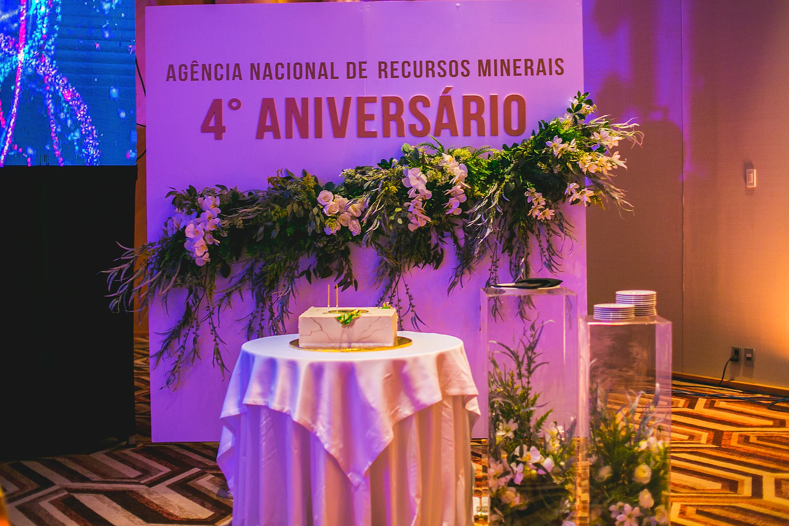 Palco do 4.º aniversário da Agência Nacional de Recursos Minerais, com bolo e arranjos de flores