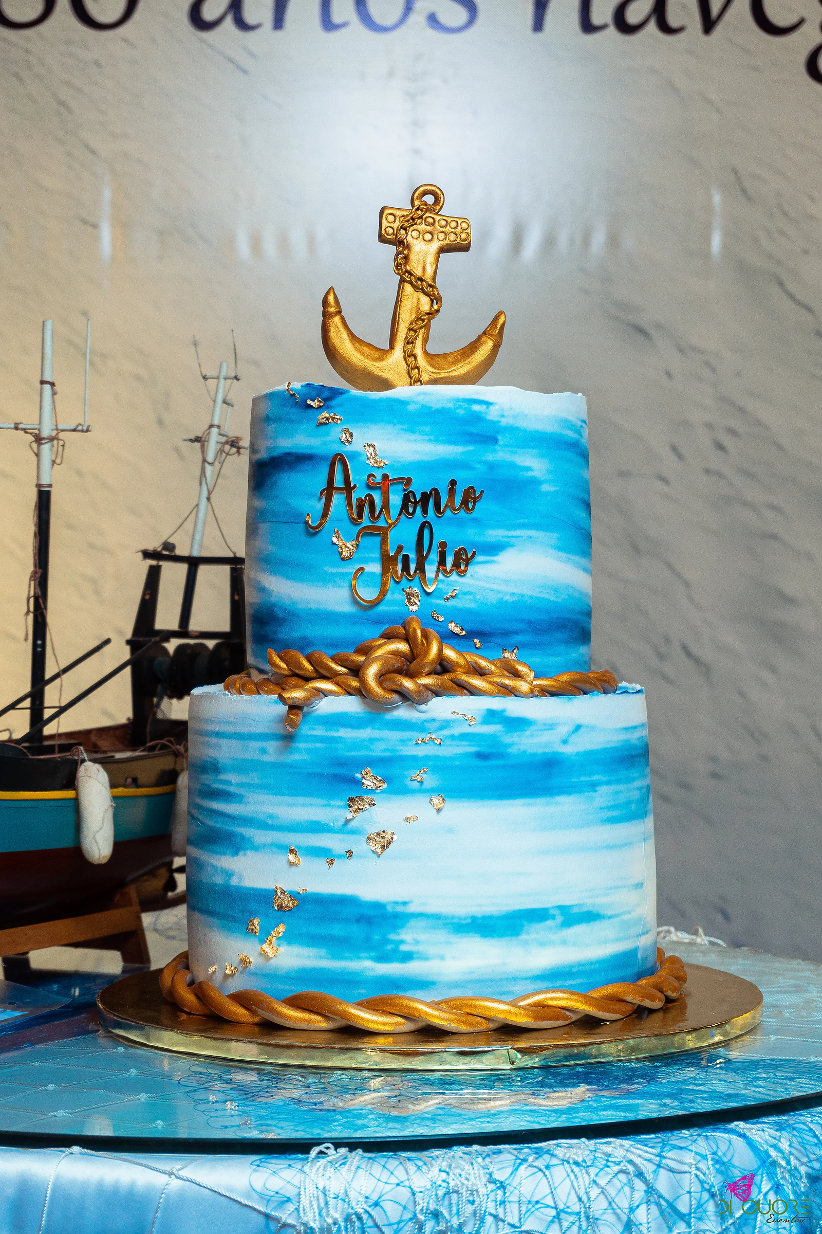 Two-tier cake in blue watercolour with a gold anchor, gold rope and the name António Júlio