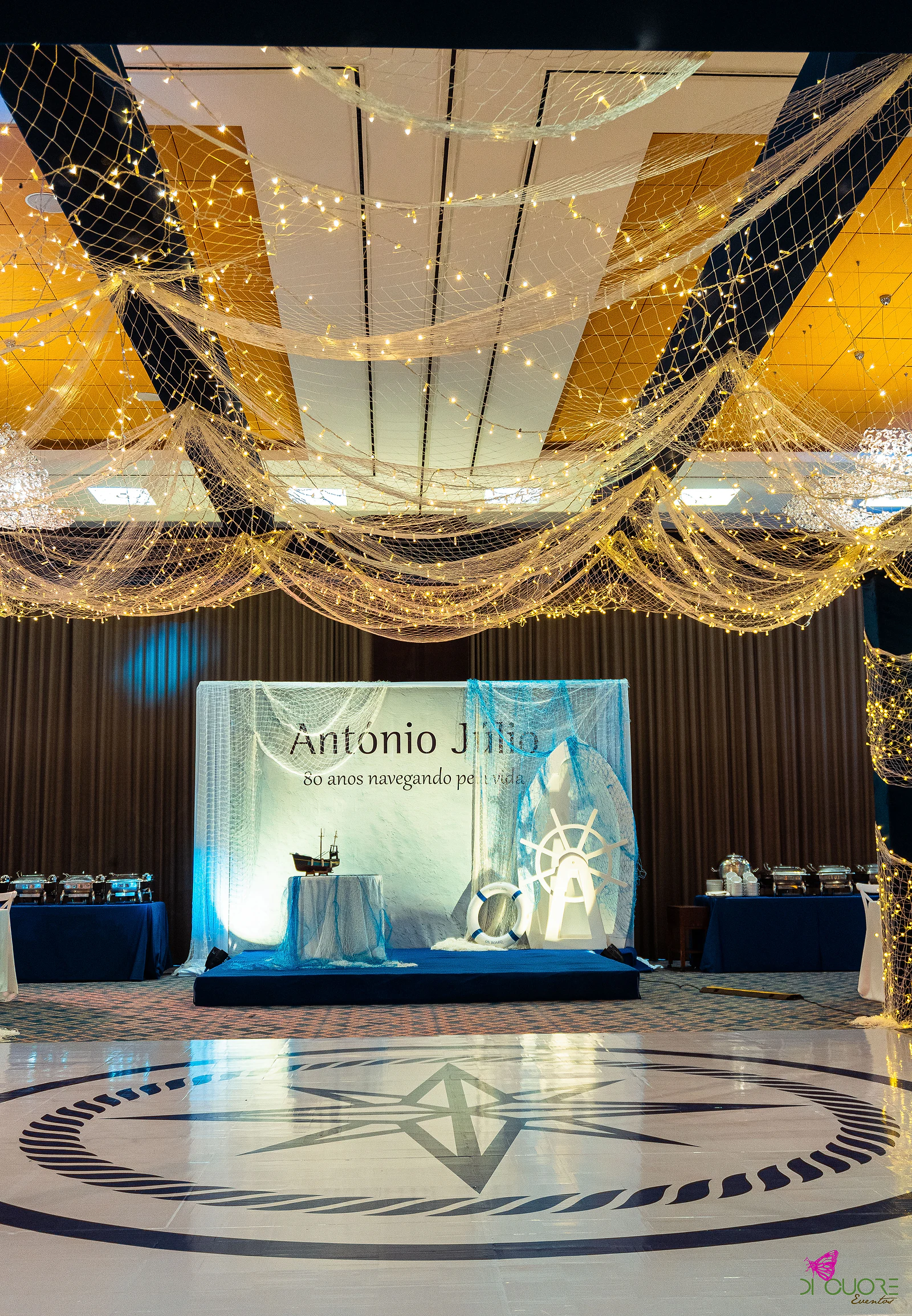 Dance floor with a compass rose beneath a fishing net strung with lights and, beyond, the backdrop reading António Júlio, 80 anos navegando pela vida