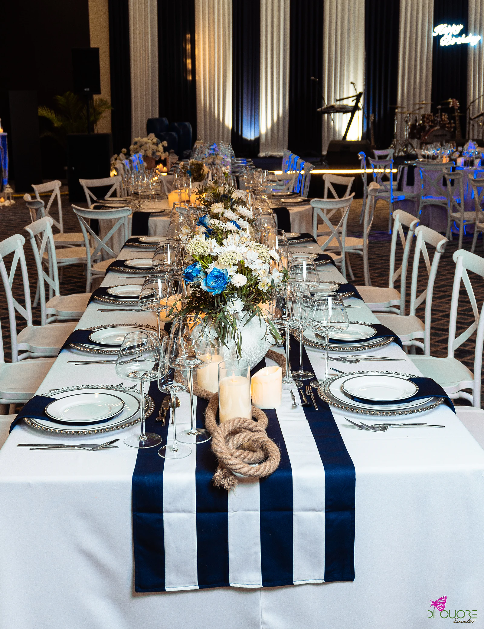 Long table with a blue and white striped runner, knotted rope, candles, white and blue flowers and white cross-back chairs