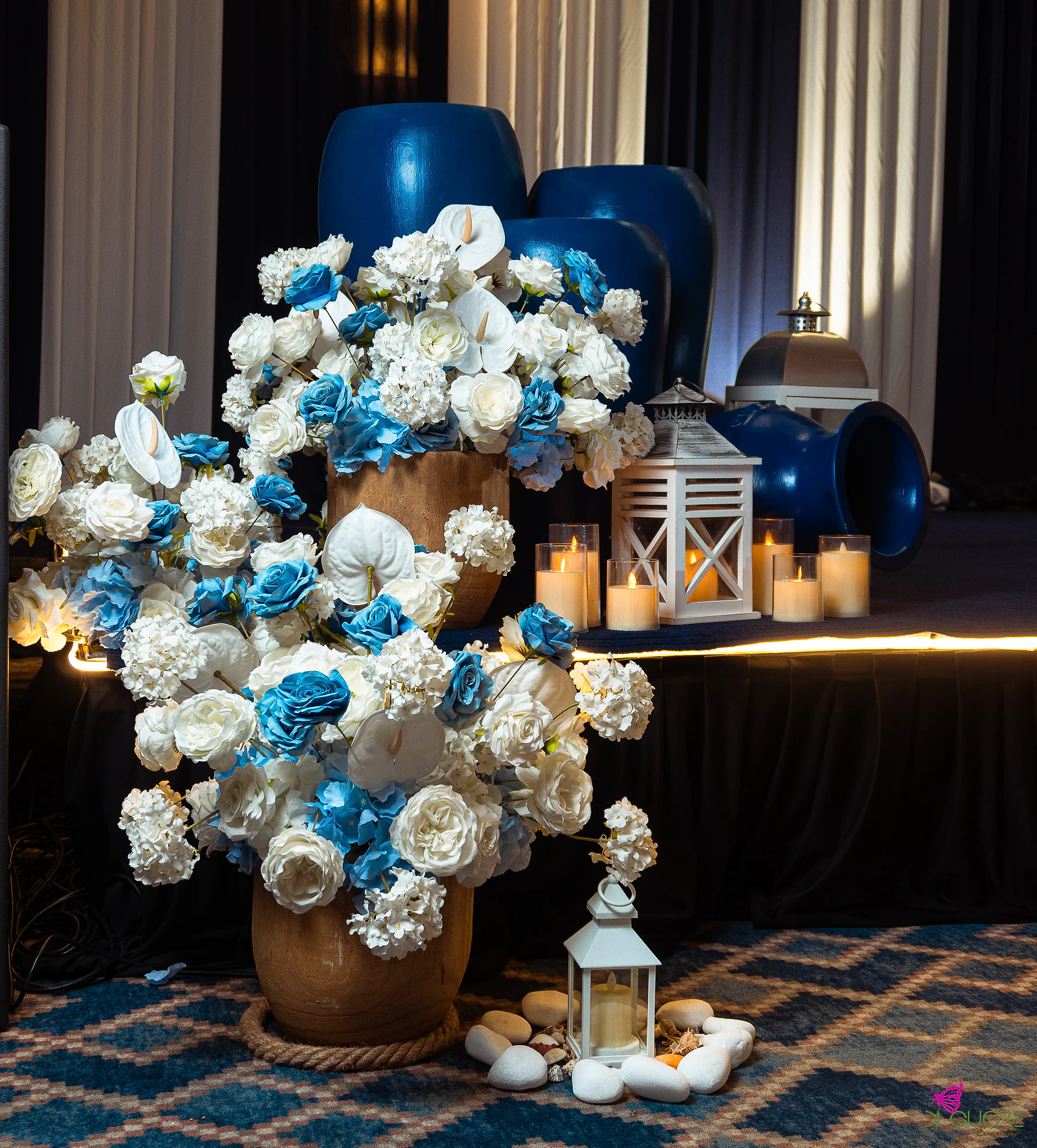 Large arrangement of blue and white roses, anthuriums and hydrangeas in a wooden pot, with lanterns, candles and big blue jars behind