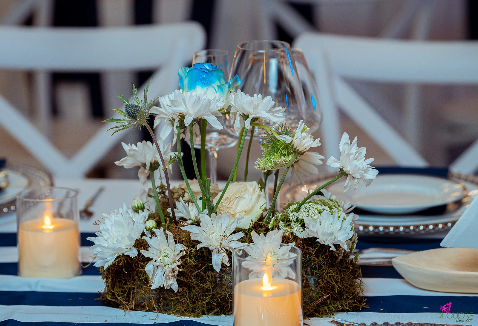 Moss centrepiece with white daisies and a blue rose, between candle glasses on the striped runner