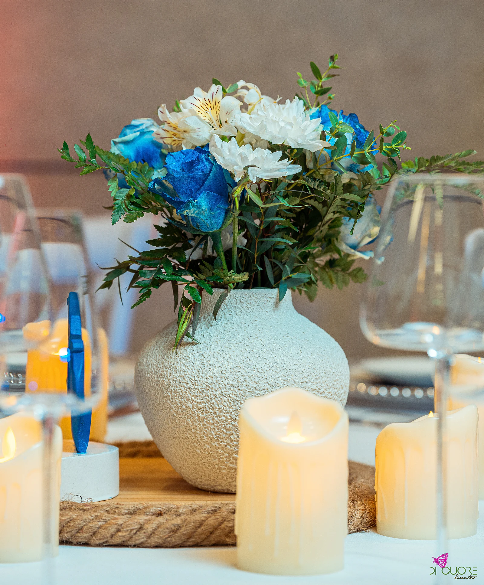 Textured white vase of blue roses and white flowers on coiled rope, between candles