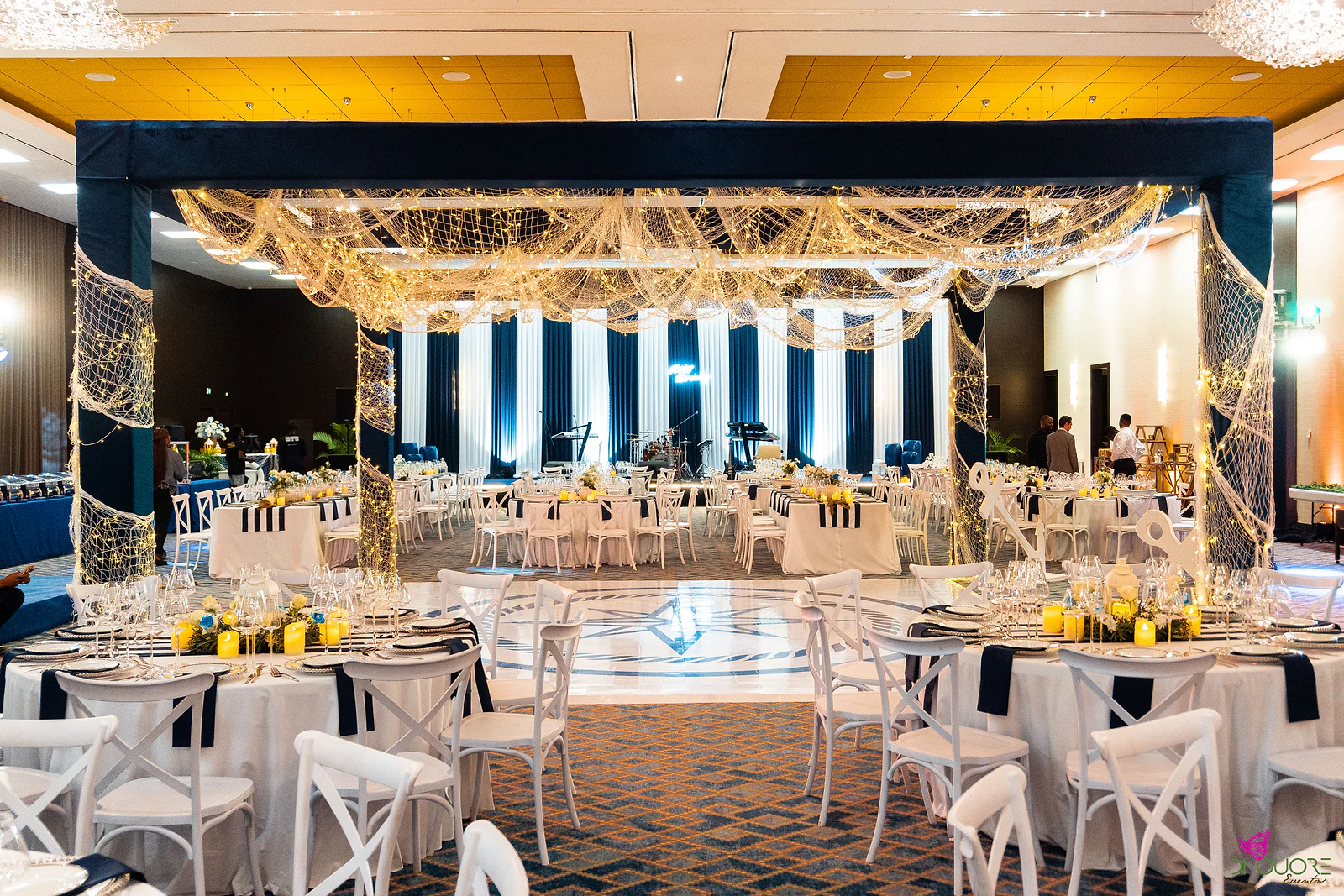 Hall with a navy fabric frame hung with nets of lights over the dance floor, laid round tables and the stage with striped curtains beyond