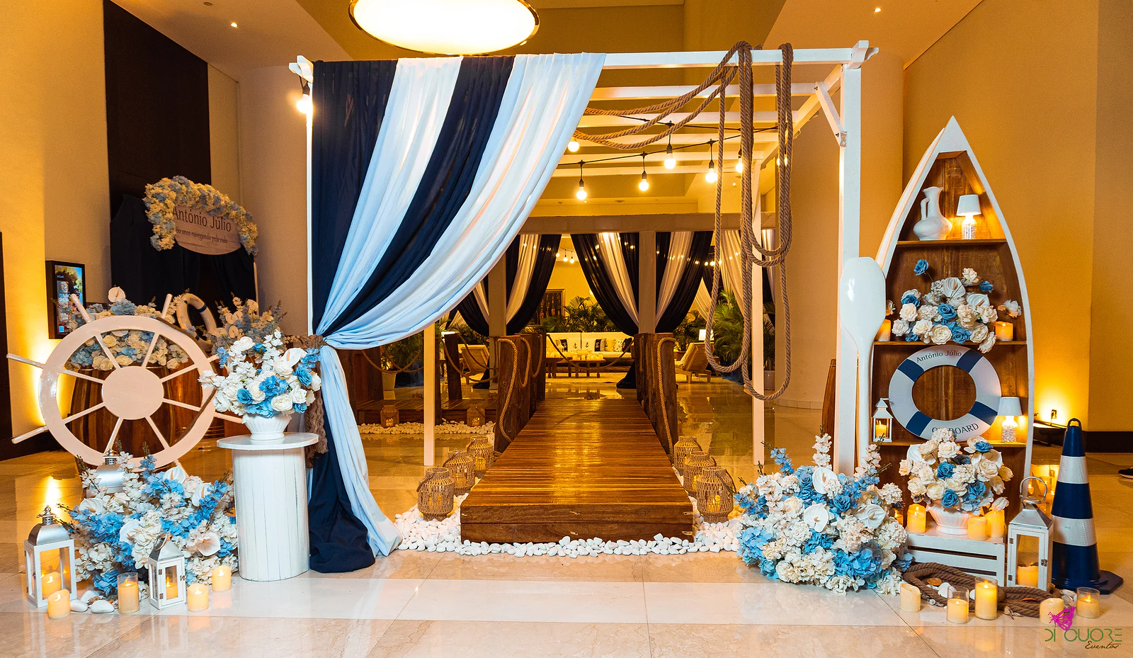 Entrance with a navy and white striped drape tied back with rope, a white ship’s wheel among flowers, a boat-shaped shelf with a lifebuoy and candles, and the wooden jetty beyond