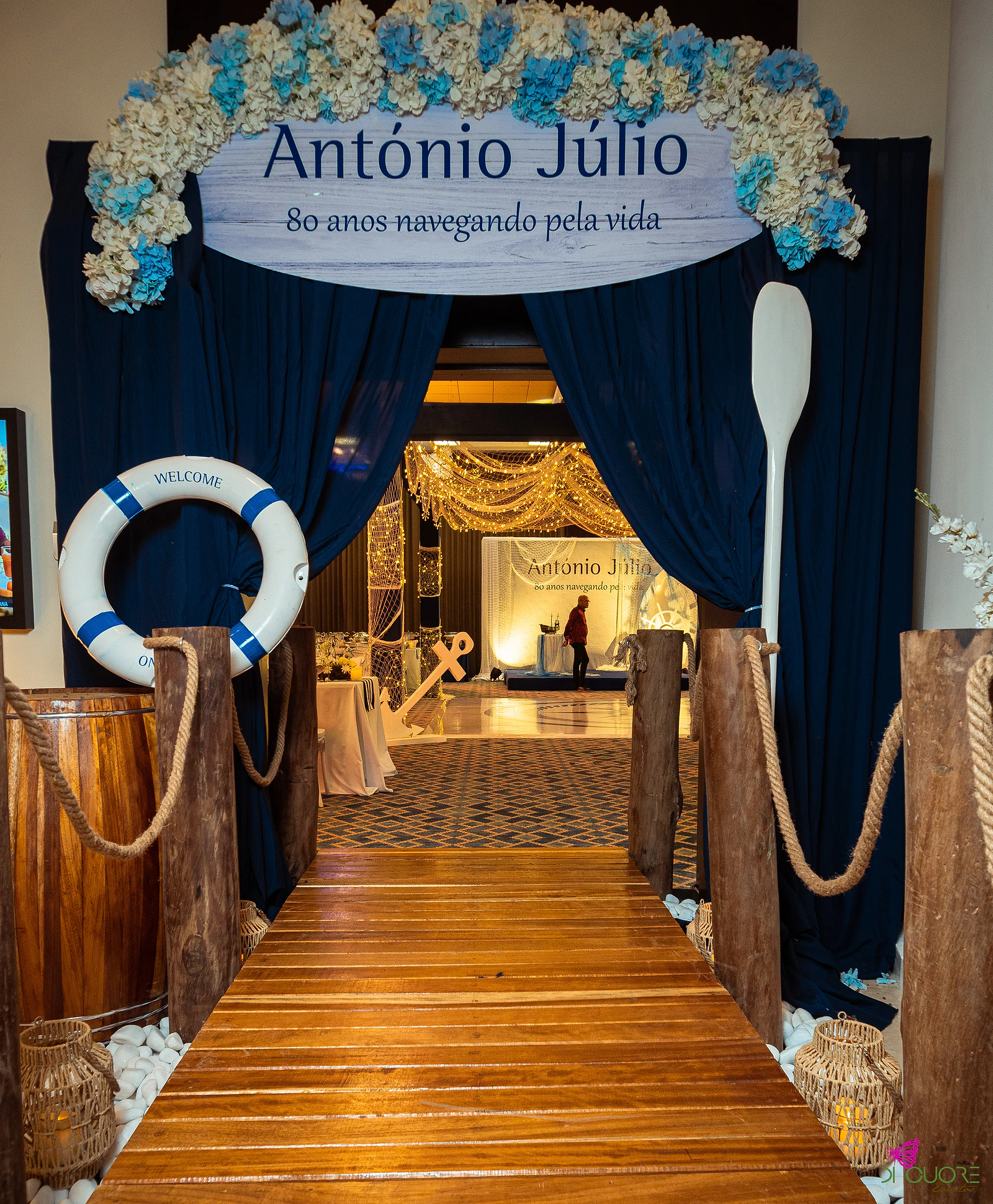 Entrance with a plank jetty, navy drapes, a lifebuoy and an oval sign topped with flowers reading António Júlio, 80 anos navegando pela vida