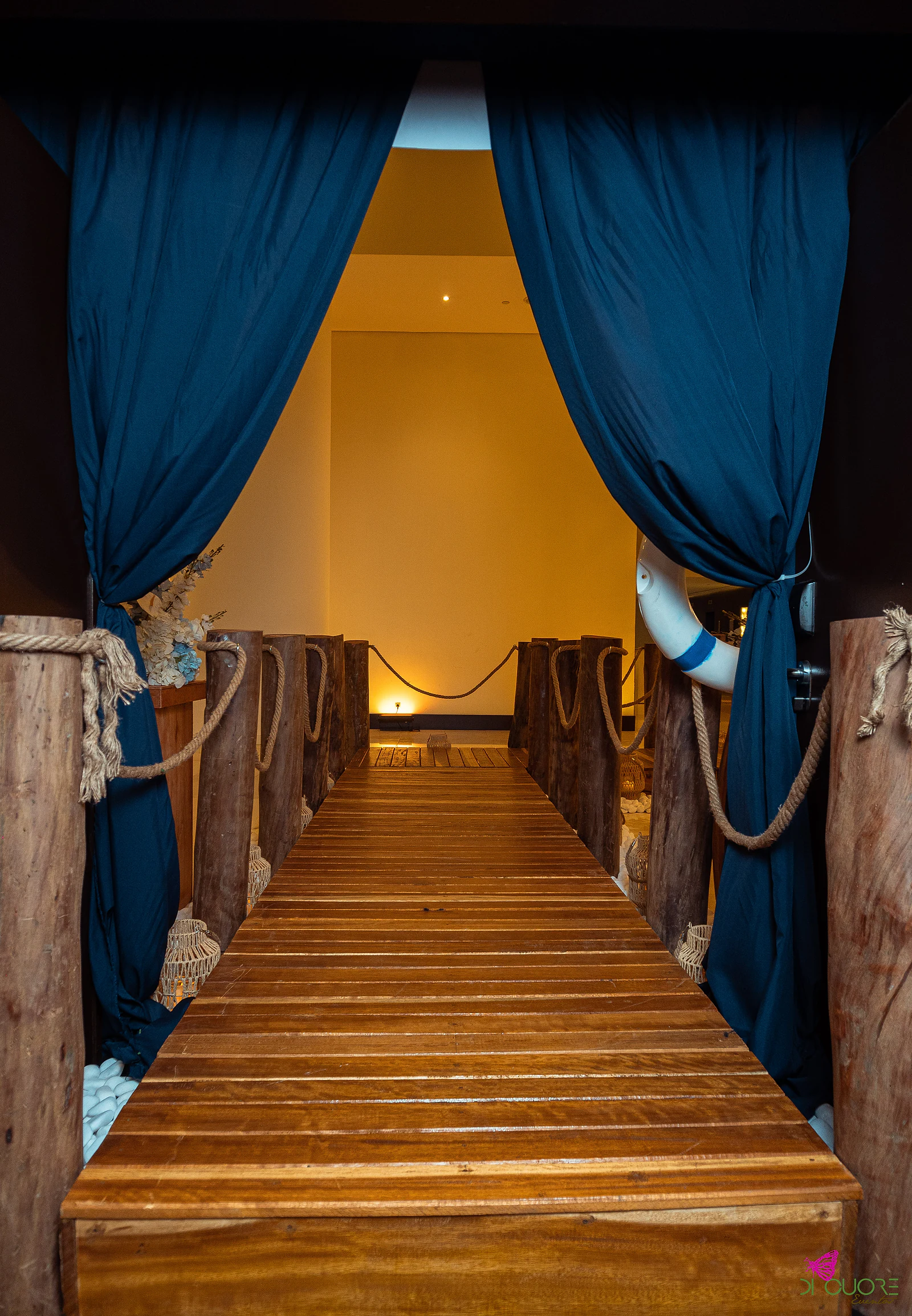 A wooden plank jetty with ropes and posts opening between two navy drapes