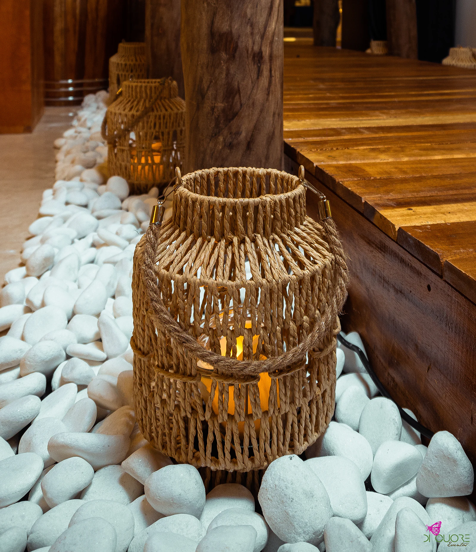 Woven rope lantern with a lit candle, among white pebbles beside the wooden jetty
