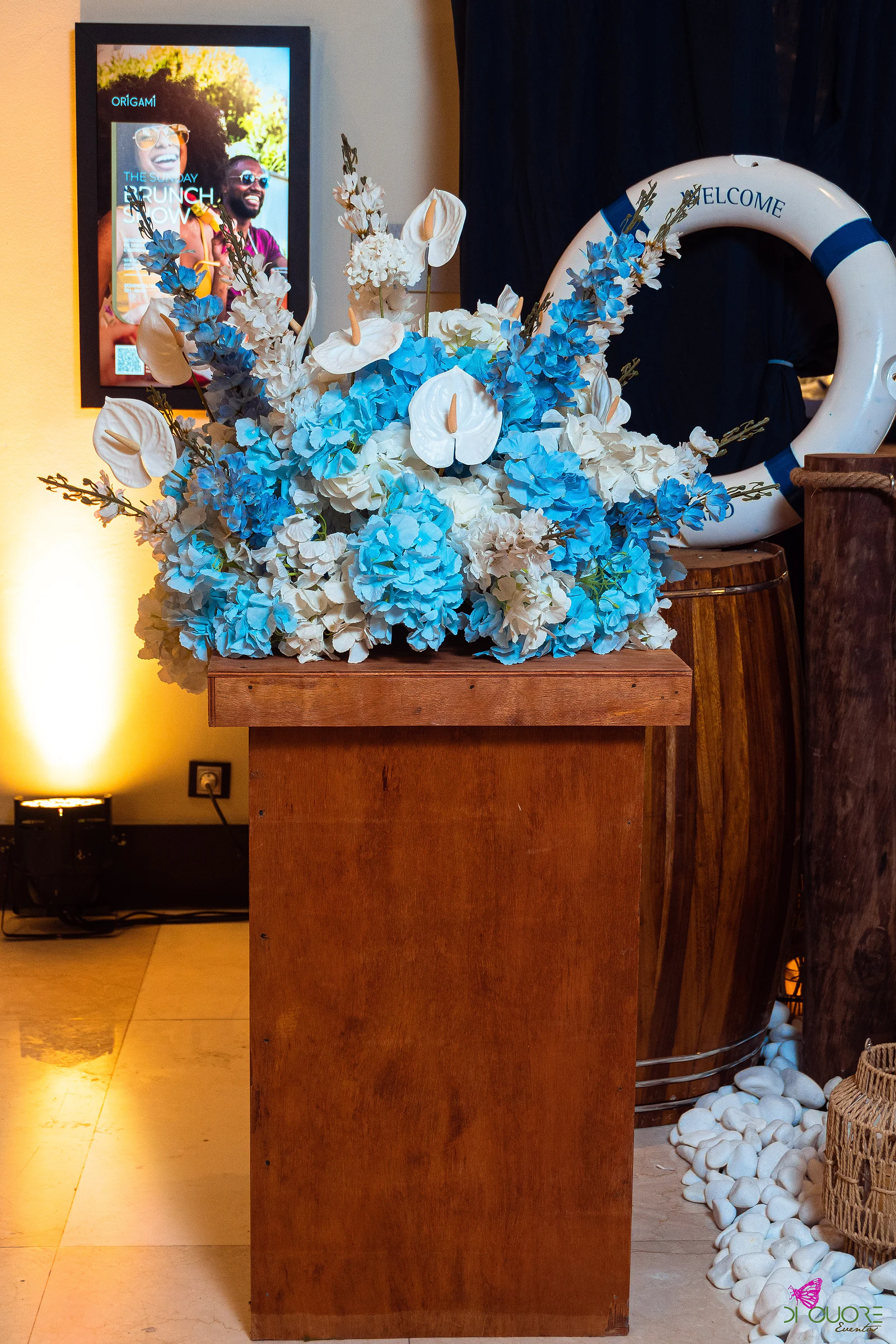 Wooden pedestal with an arrangement of blue and white flowers and anthuriums, beside a barrel and a lifebuoy marked Welcome