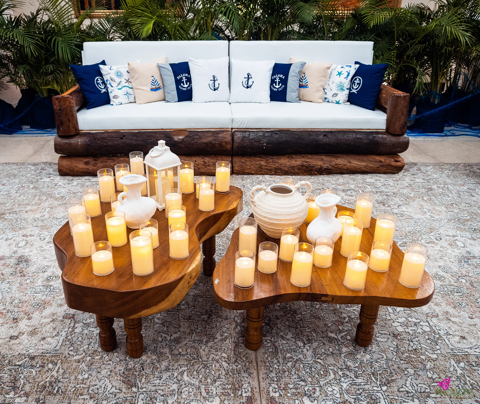 White sofa with anchor-embroidered cushions on a log frame, before two low wooden tables with candles and white jugs
