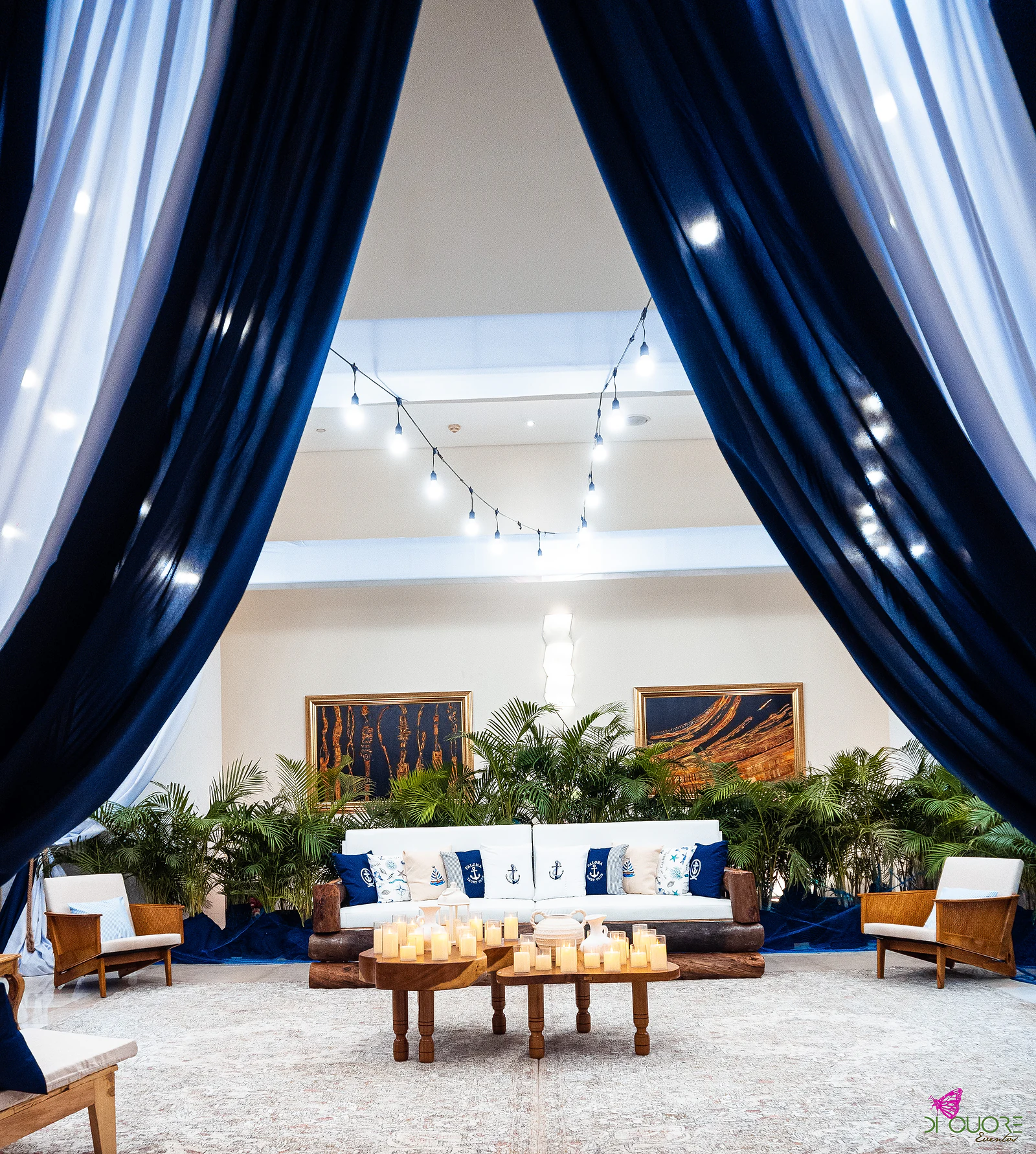 Lounge seen between navy and white drapes, with a string of lights, a white sofa with anchor cushions, armchairs and low tables with candles