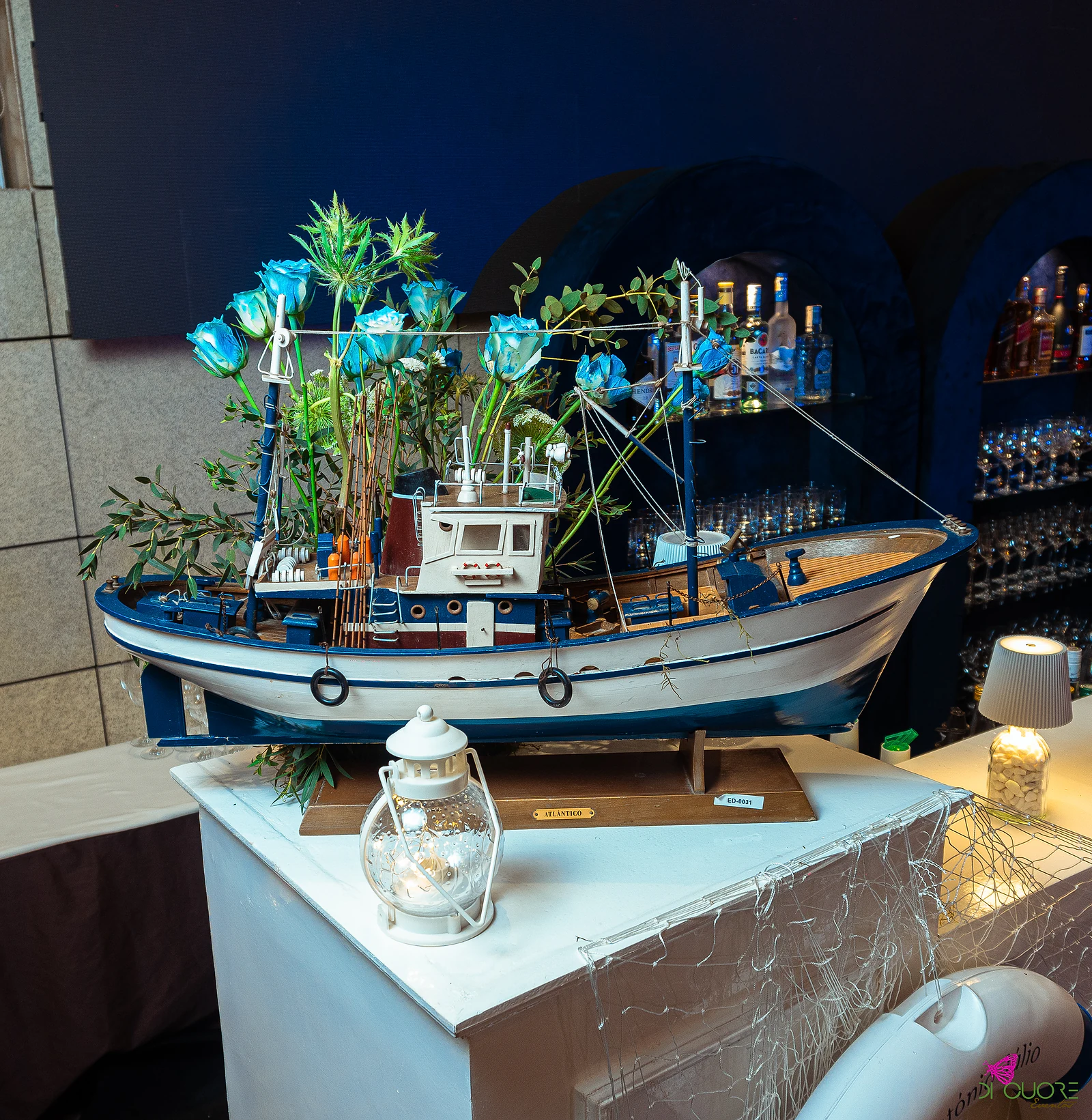 A miniature fishing boat with blue roses and foliage rising from the deck, on a white plinth with a lantern and fishing net, before the bar