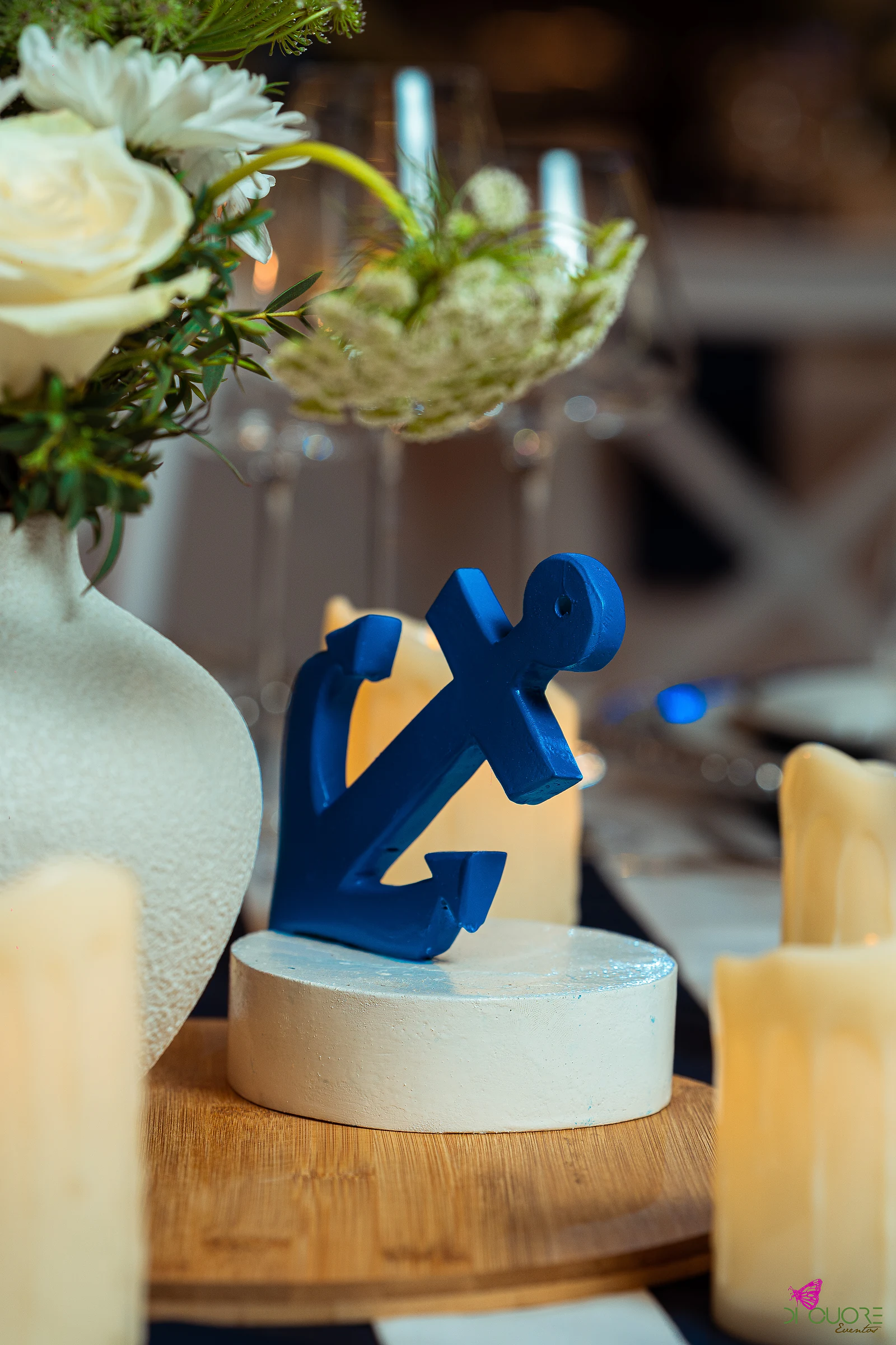 A blue anchor on a round white stand, between candles and a vase of white flowers on a laid table