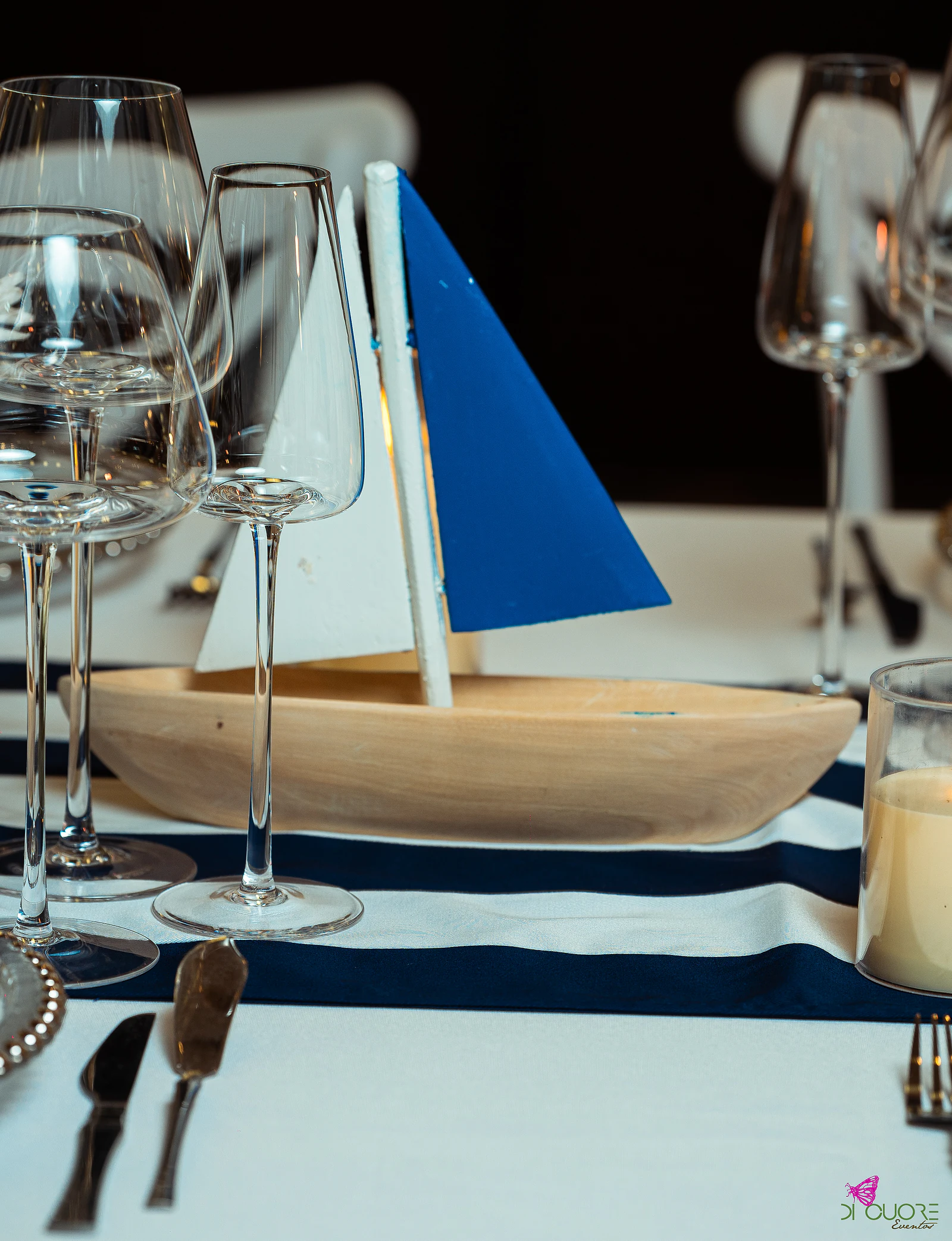 Pale wooden sailing boat with one white and one blue sail on the blue and white striped runner, among glasses and a candle