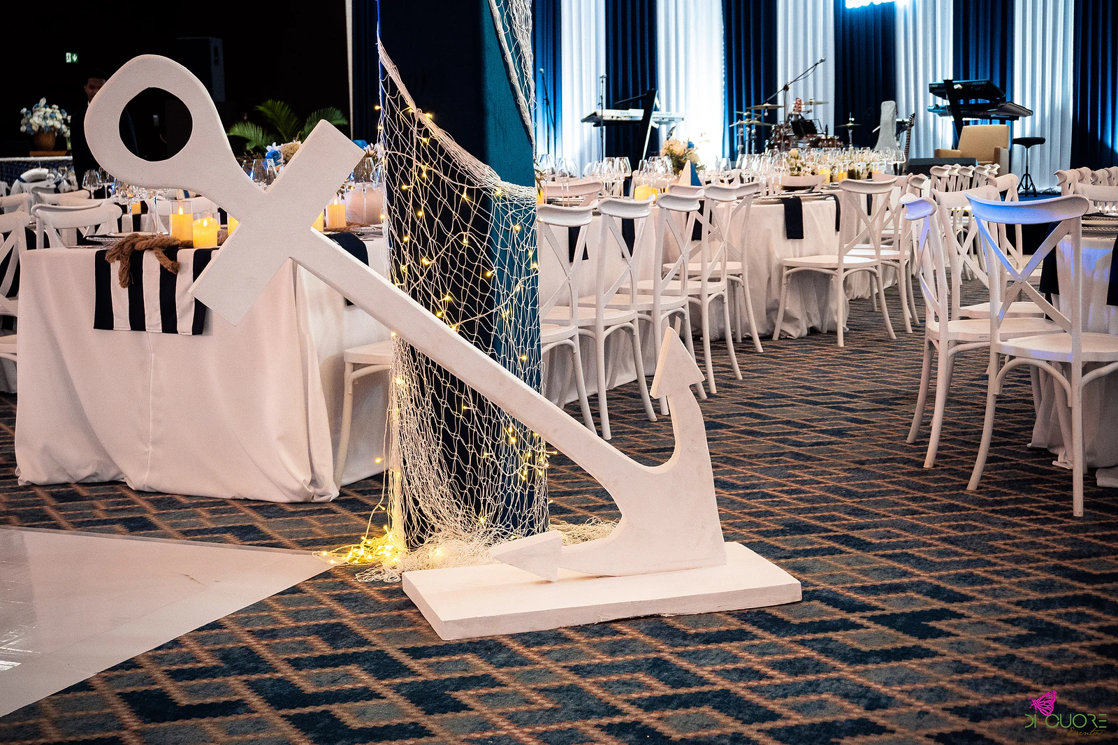 Large white anchor on a base, beside a column wrapped in netting with fairy lights, next to the dance floor and laid tables, with the stage beyond