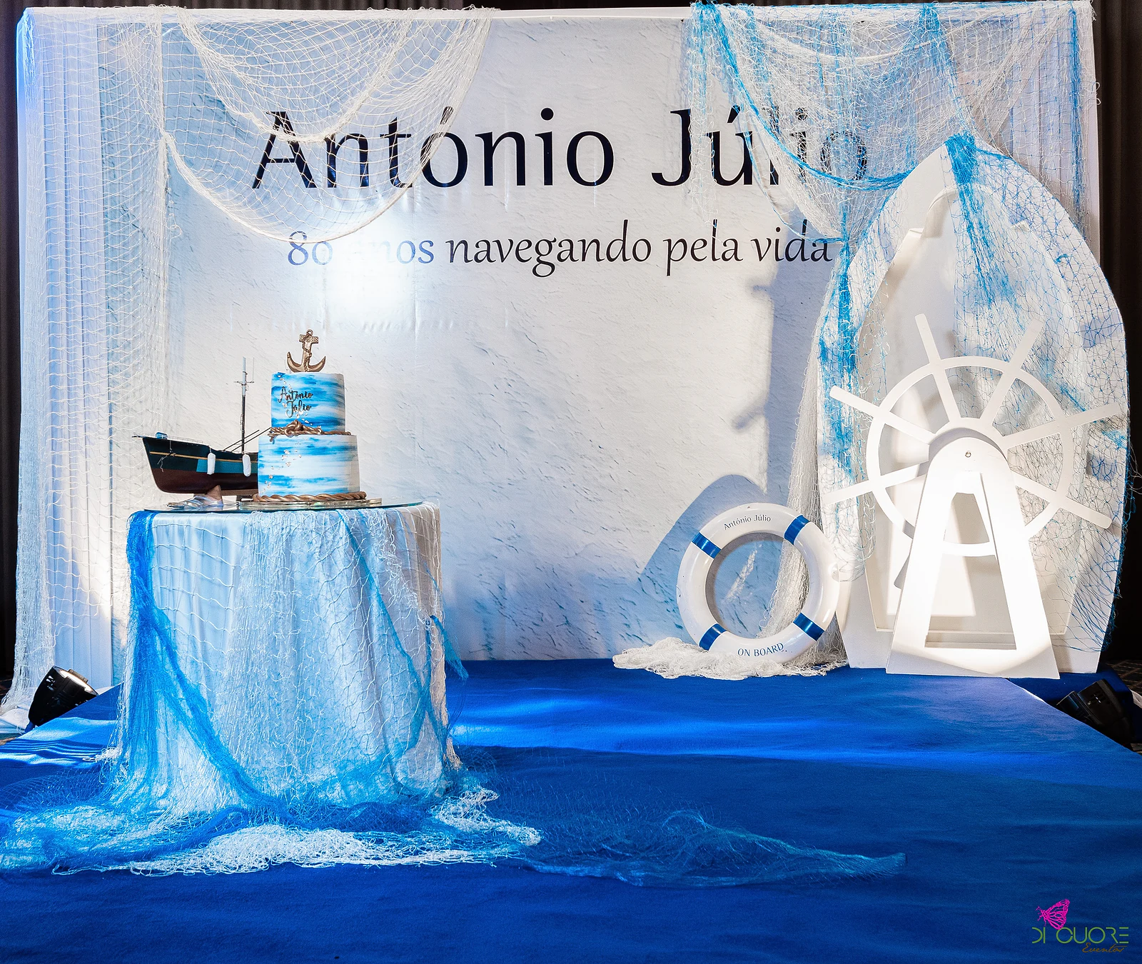 Backdrop reading António Júlio, 80 anos navegando pela vida, a blue cake with a gold anchor and a fishing boat on a net-covered table, with a white lifebuoy and ship’s wheel