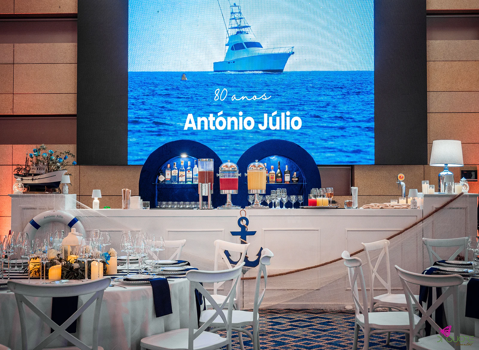 Screen showing a boat at sea with the text 80 anos António Júlio, above the white bar with blue niches, anchor, rope and netting, and laid tables in front
