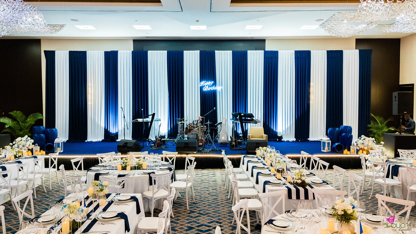 Stage with navy and white striped curtains, a Happy Birthday neon sign, drums and keyboards, and long laid tables in the foreground