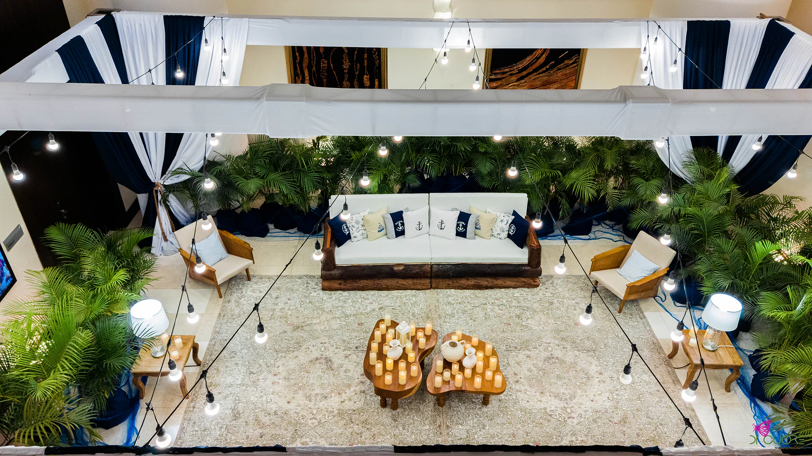 Lounge seen from above, with a white sofa and anchor cushions, armchairs, two wooden tables with candles, a pale rug, palms and a string of lights