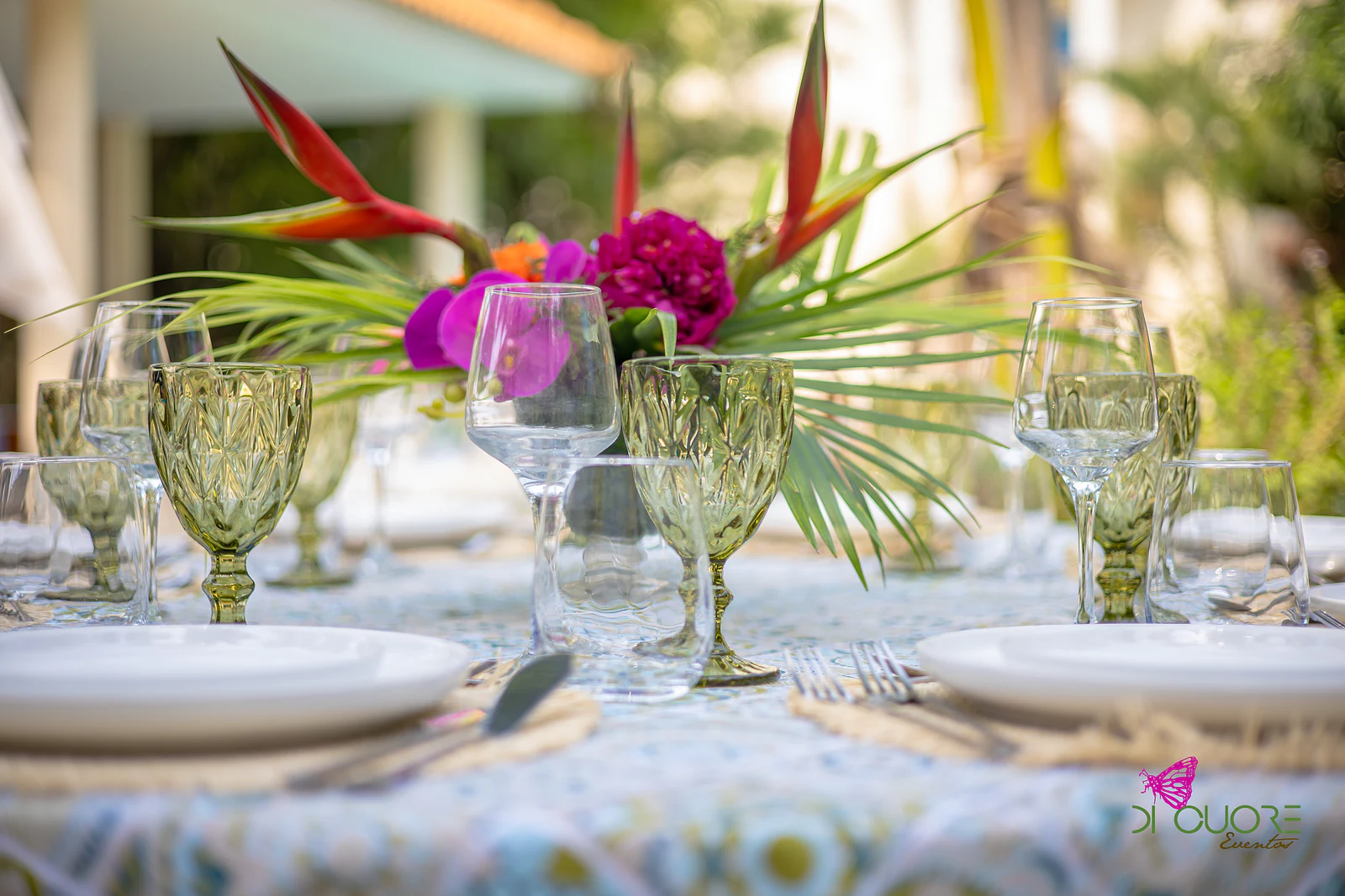 Centro de mesa com helicónia vermelha, folhas de palmeira e uma flor cor-de-rosa, entre copos verdes lapidados e pratos brancos