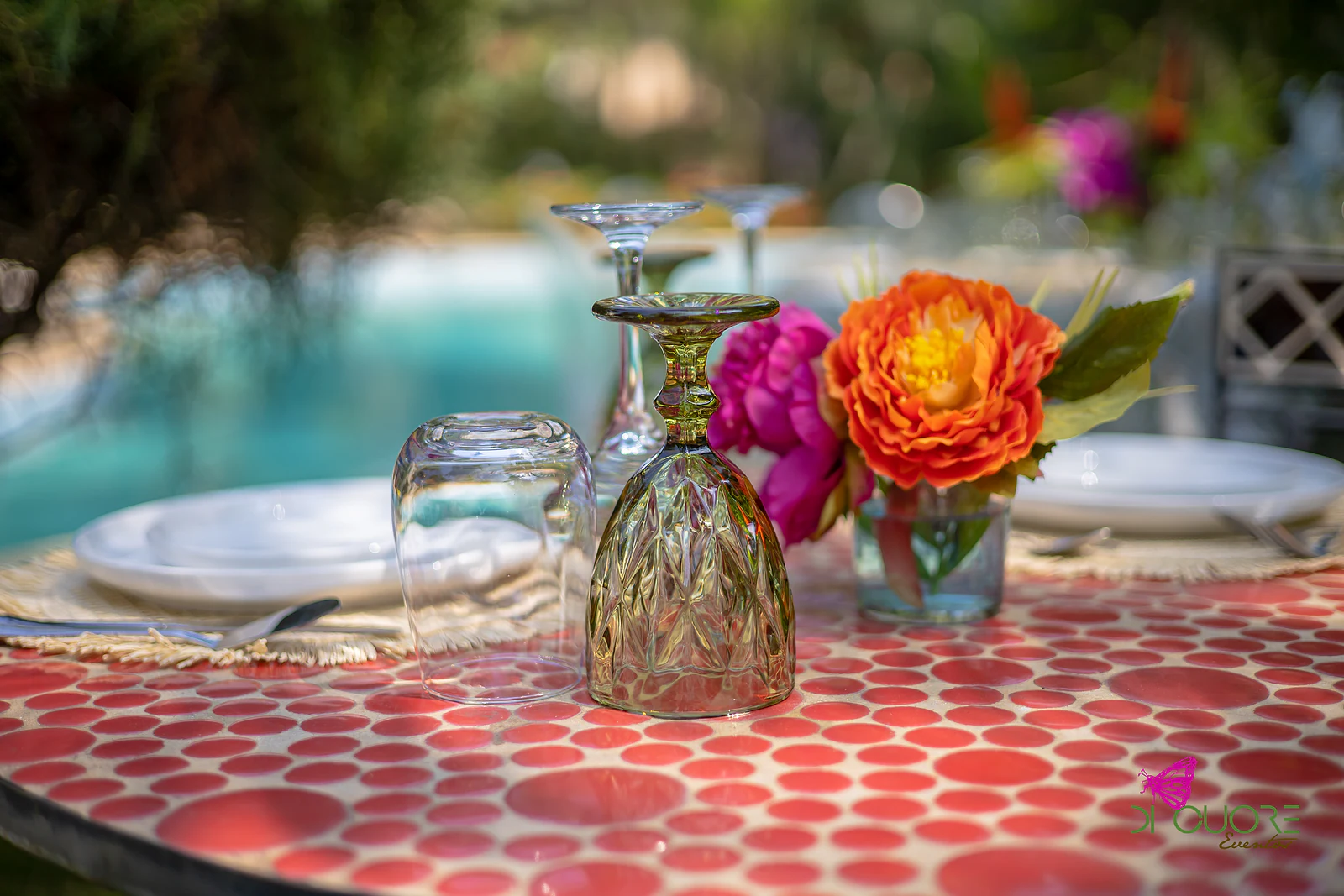 Mesa de mosaico de círculos vermelhos com um copo verde virado ao contrário e flores cor de laranja e cor-de-rosa, com a piscina desfocada ao fundo