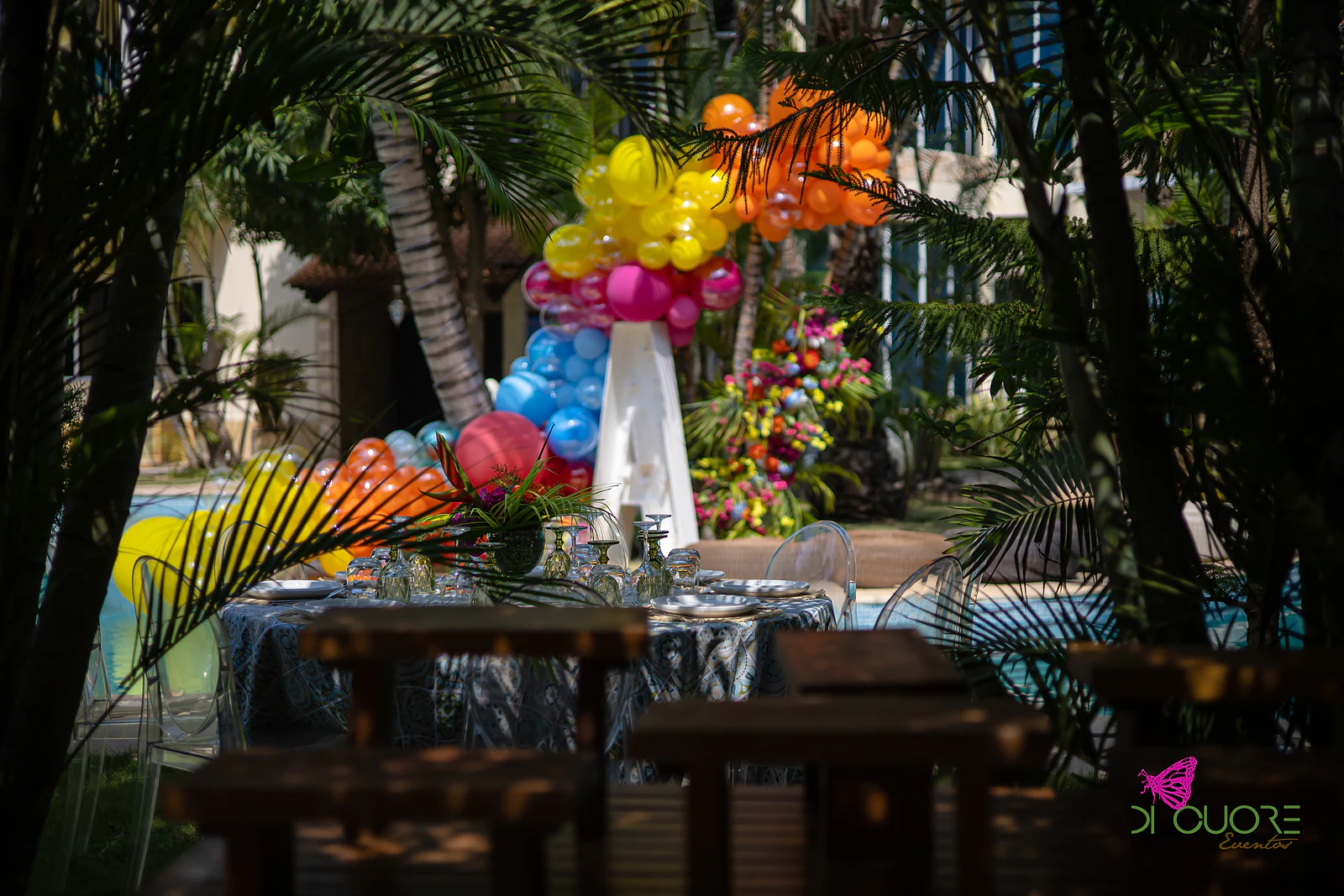 Escultura de balões amarelos, cor-de-rosa, azuis e cor-de-laranja sobre uma letra A branca, com flores tropicais, vista entre palmeiras e mesas postas em primeiro plano