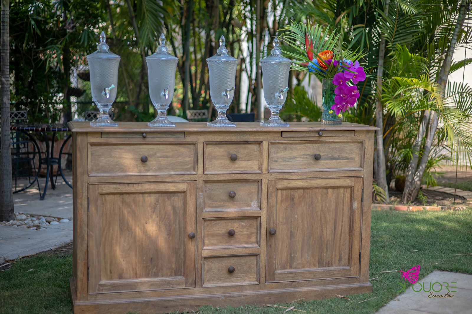 Aparador de madeira com quatro dispensadores de vidro para bebidas e um ramo de flores tropicais cor-de-rosa e laranja, no relvado