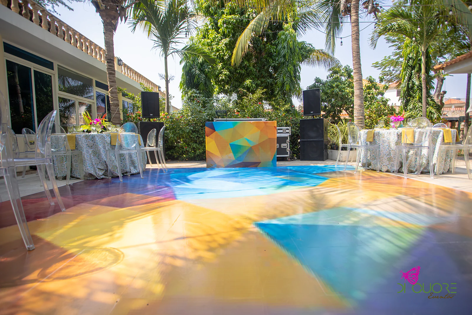 Pista de dança vista ao nível do chão, com triângulos de cor, a cabina do DJ e colunas de som entre palmeiras e mesas redondas